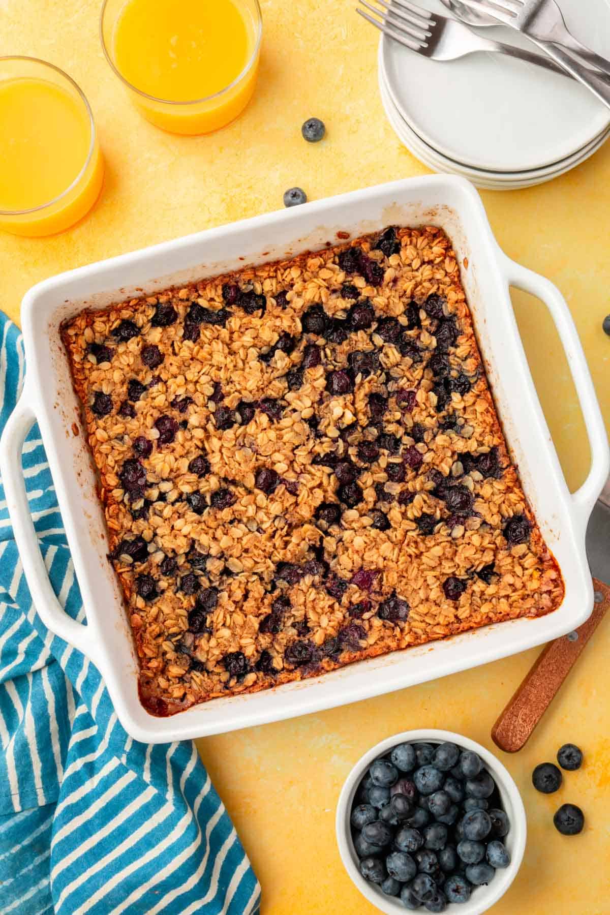 An overhead view of a baking dish with blueberry baked oatmeal in it with two glasses of orange juice and a bowl of blueberries on the side.