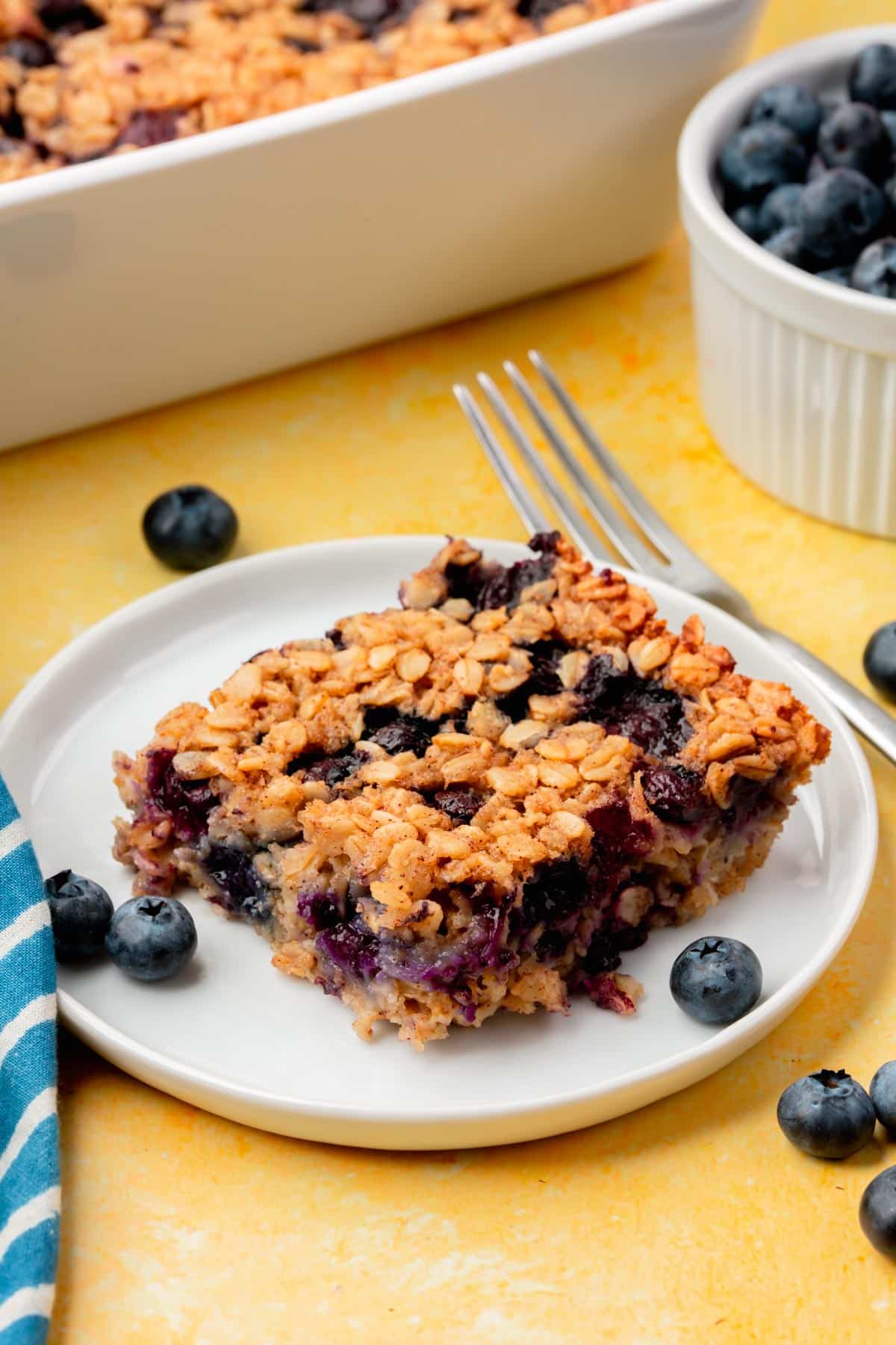 A square of blueberry baked oatmeal on a small plate with a bowl of blueberries and a baking dish of baked oatmeal in the background.