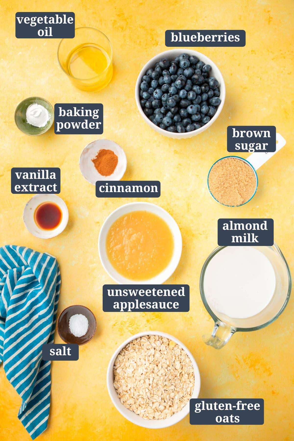 Small bowls of ingredients on a yellow table to make blueberry baked oatmeal, including vegetable oil, baking powder, vanilla extract, brown sugar, unsweetened applesauce, almond milk, cinnamon, and gluten-free oats.