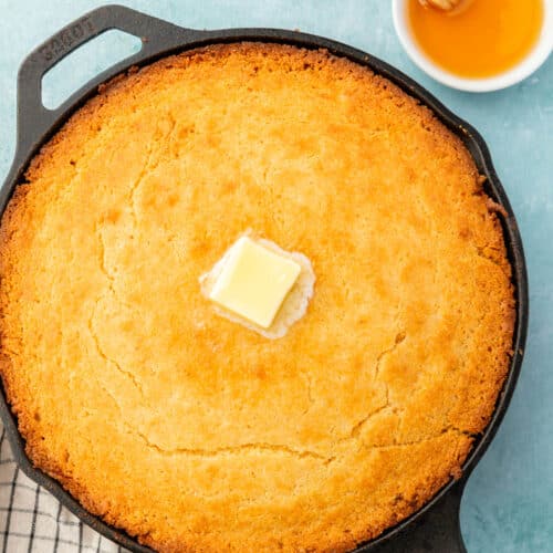 A cast iron skillet filled with gluten-free cornbread topped with a pat of melted butter with a ramekin of honey and a plate of butter on the side.