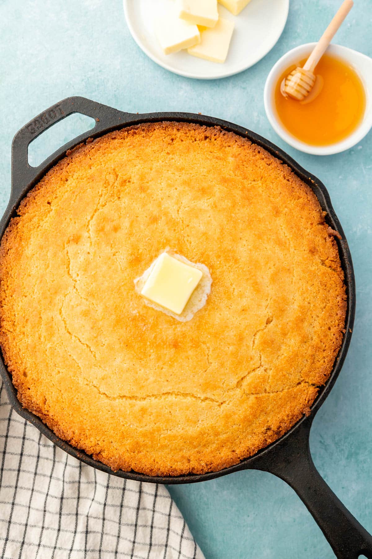 A cast iron skillet filled with gluten-free cornbread topped with a pat of melted butter with a ramekin of honey and a plate of butter on the side.