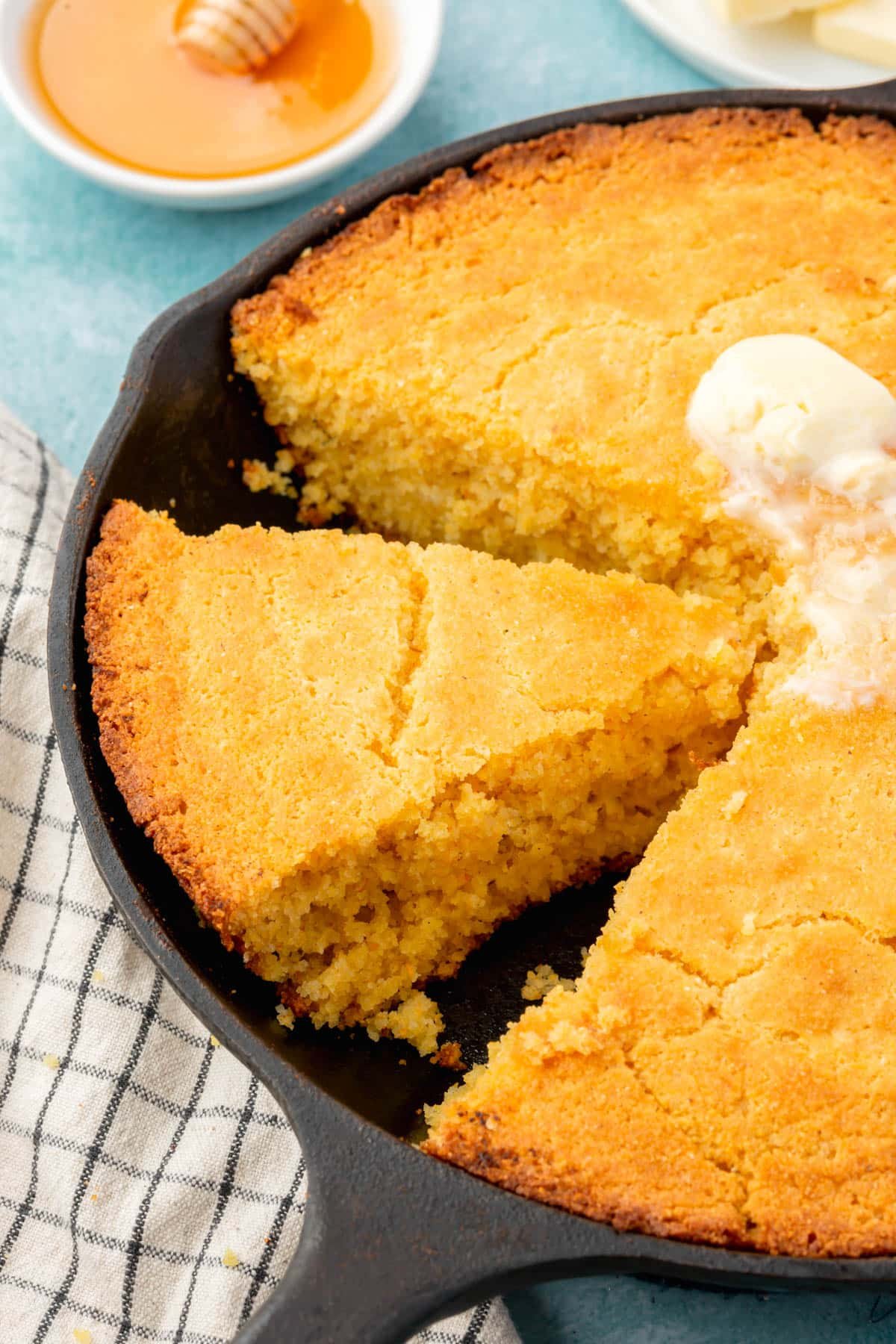 A closeup of slices of gluten-free cornbread in a cast iron skillet with melted butter on top and a ramekin of honey in the background.