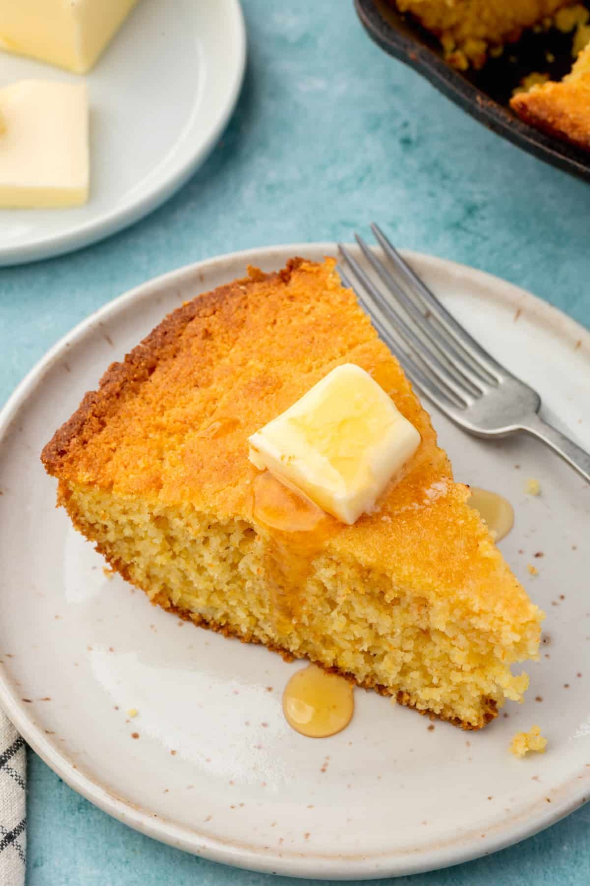 A wedge of gluten-free cornbread topped with melted butter and honey on an appetizer plate with a fork.