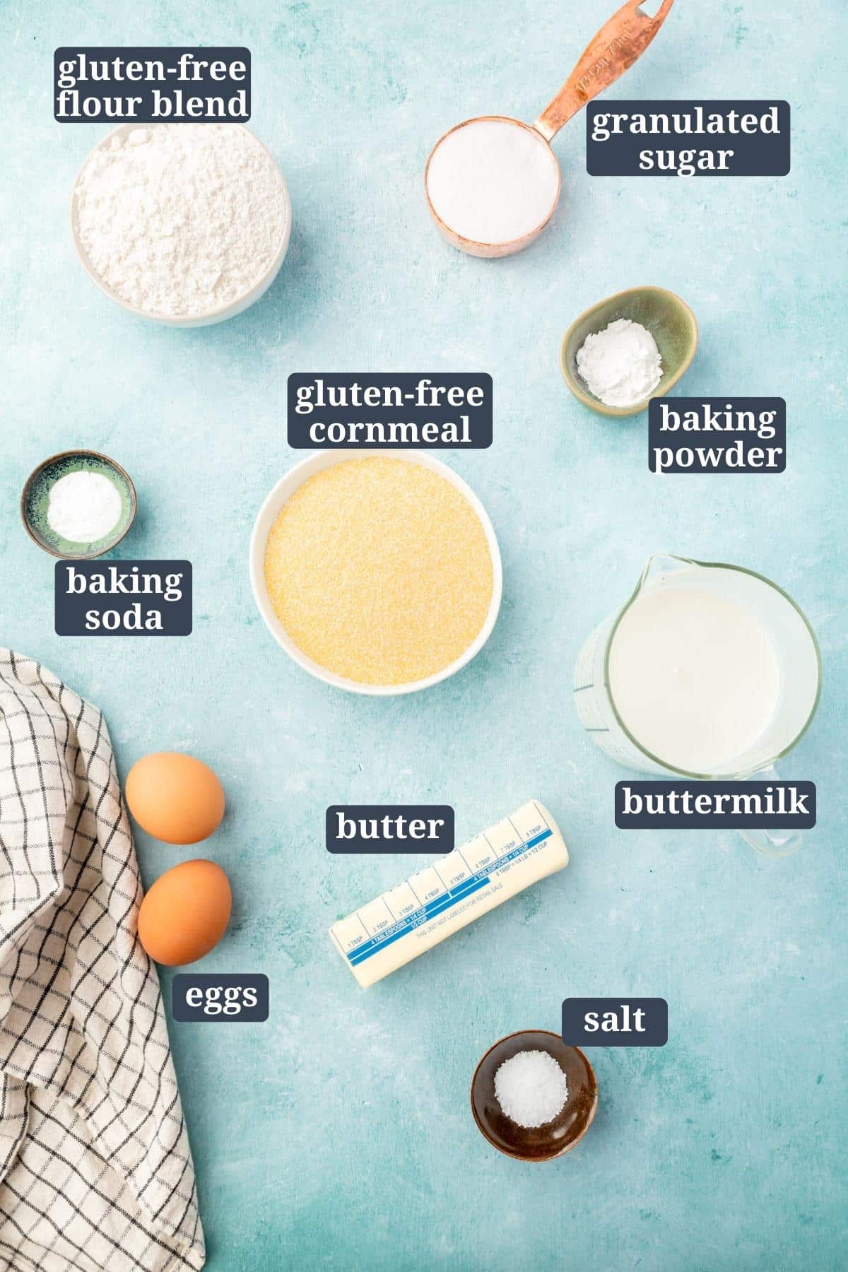An overhead view of ingredients in small bowls on a blue table for making gluten-free cornbread, including gluten-free flour, sugar, salt, baking soda, baking powder, buttermilk, cornmeal, eggs, butter with text overlays over each ingredient.
