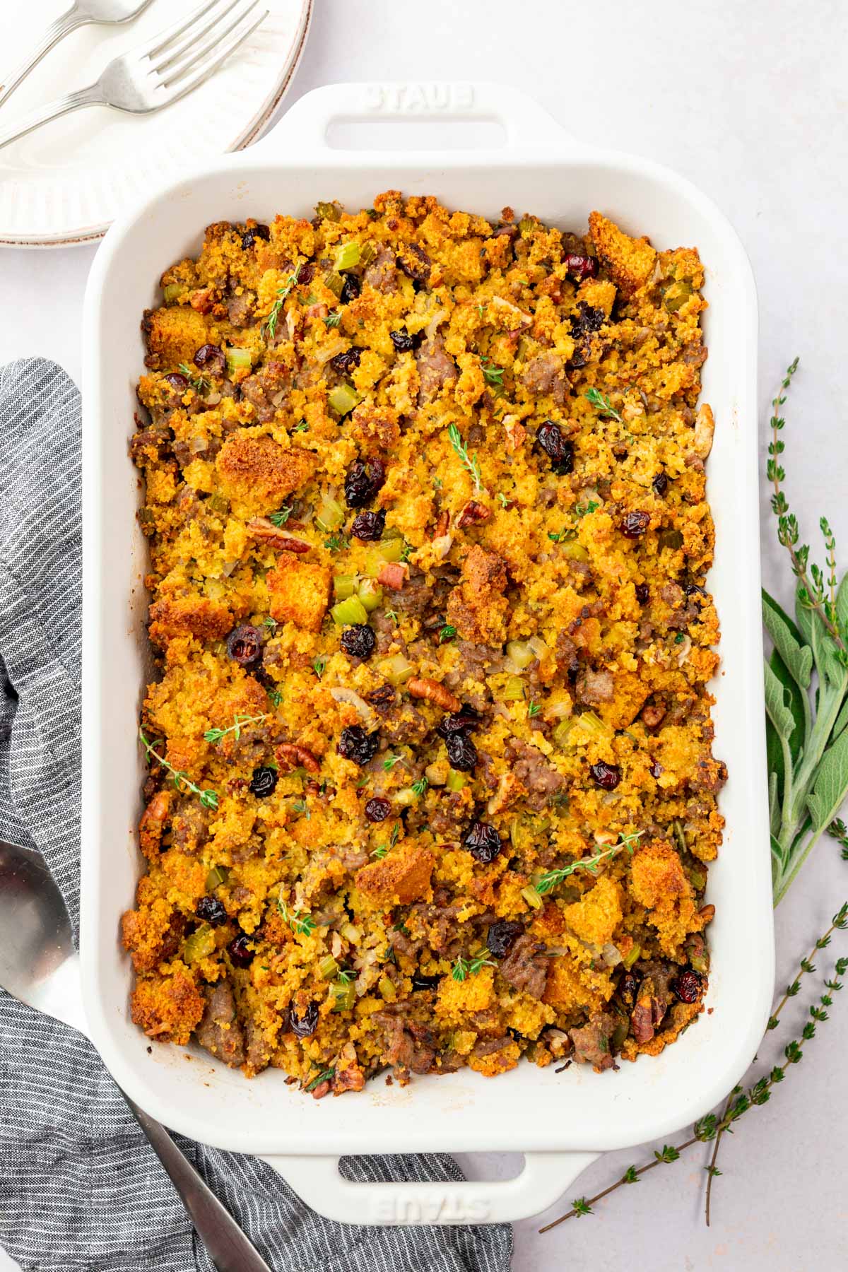 An overhead view of a rectangle casserole dish filled with gluten-free cornbread stuffing topped with fresh thyme, dried cranberries, and Italian sausage.