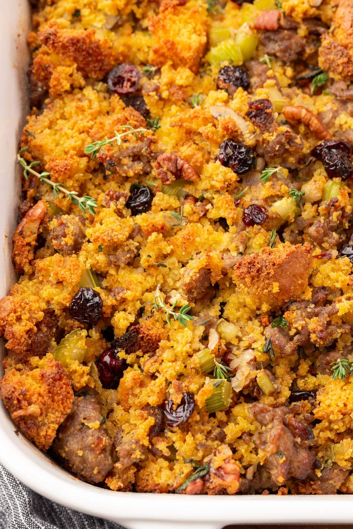 A closeup of a casserole dish with gluten-free cornbread sausage stopping topped with fresh thyme and dried cranberries.