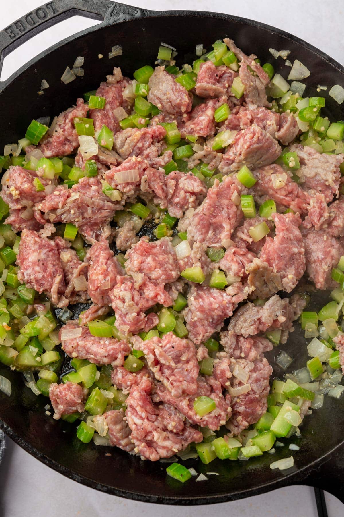 A cast iron skillet filled with raw Italian sausage mixed together with cooked diced celery and diced onions.