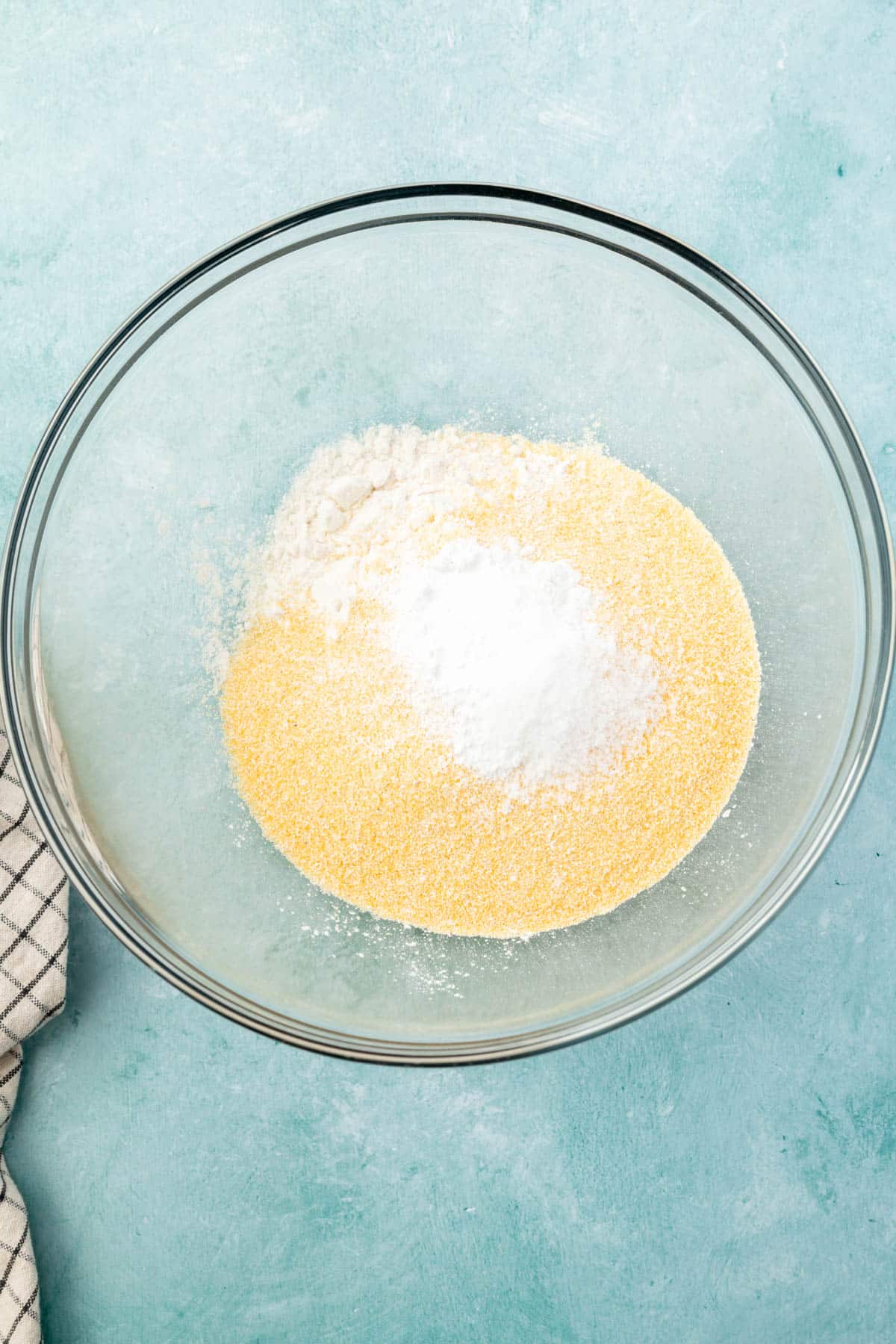 A glass mixing bowl with cornmeal, gluten-free flour, salt, baking soda, and baking powder before mixing.