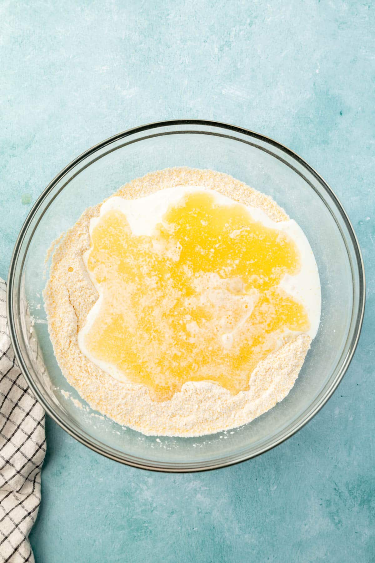 A glass mixing bowl with dry ingredients for cornmeal topped with buttermilk, whisked eggs, and melted butter.