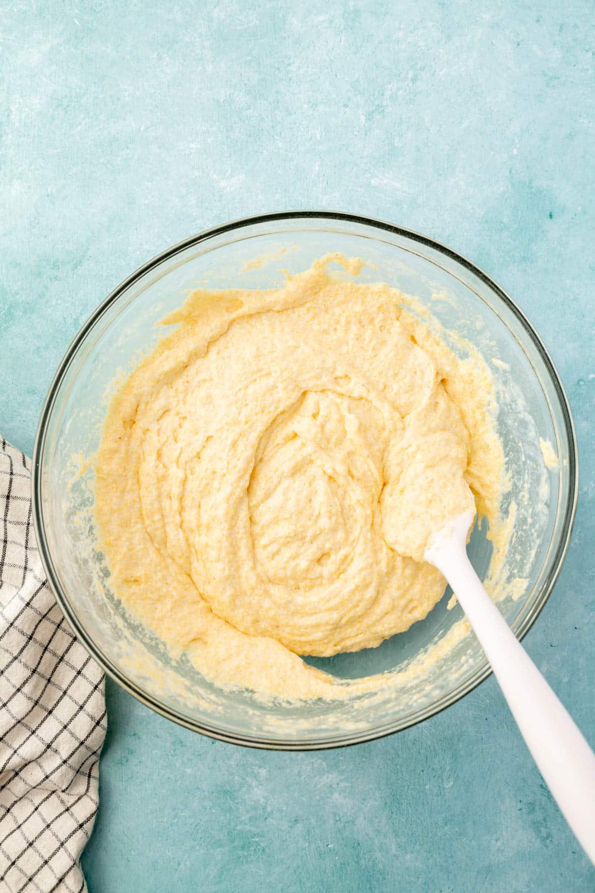 A glass mixing bowl with cornbread batter that is being mixed up with a spatula.