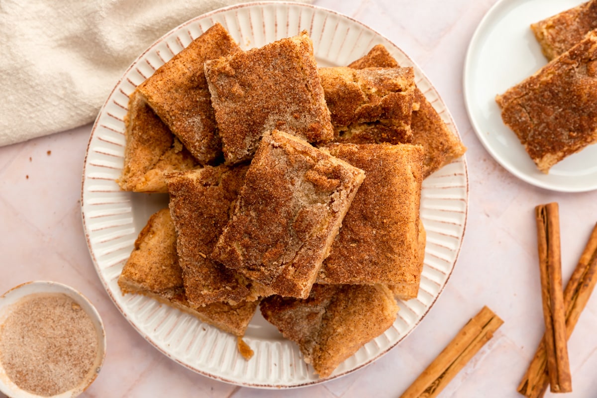 A pile of snickerdoodle blondies on a plate with cinnamon sticks and cinnamon sugar next to the plate.