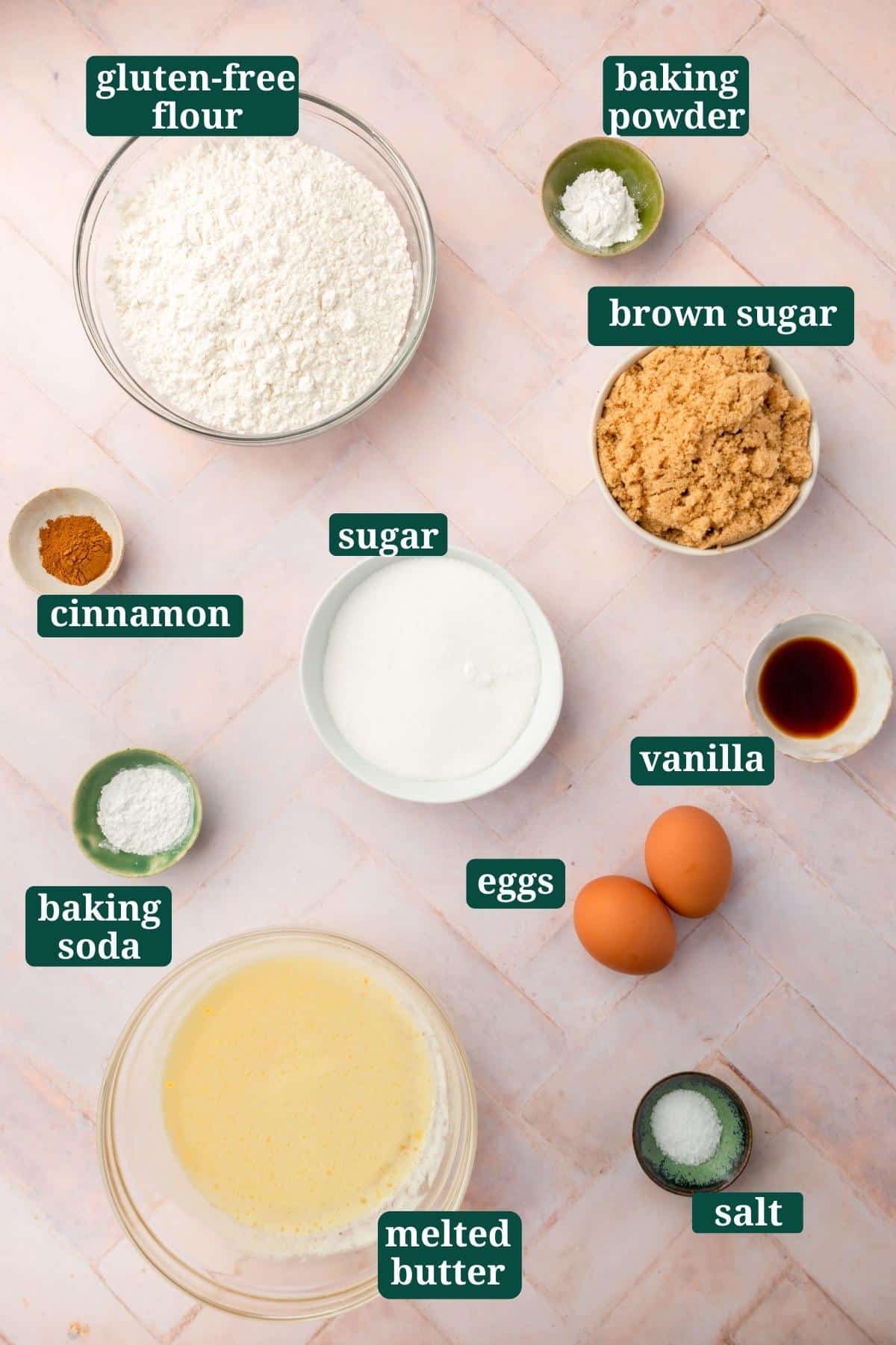 Small bowls of ingredients on a pink table to make gluten-free snickerdoodle bars, including gluten-free flour, baking powder, brown sugar, vanilla, sugar, cinnamon, baking soda, eggs, melted butter and salt with text overlays over each ingredient.