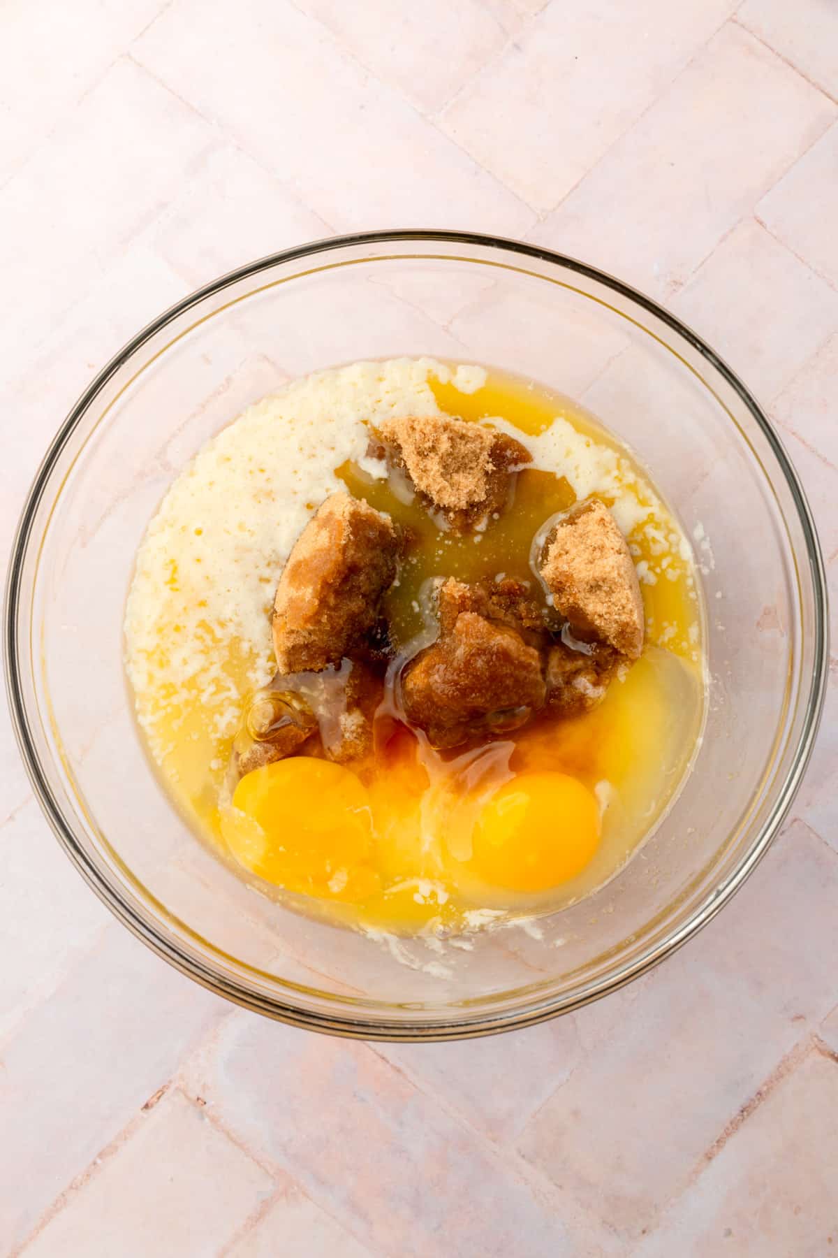 A glass mixing bowl with eggs, brown sugar, melted butter, vanilla, and granulated sugar before mixing together.