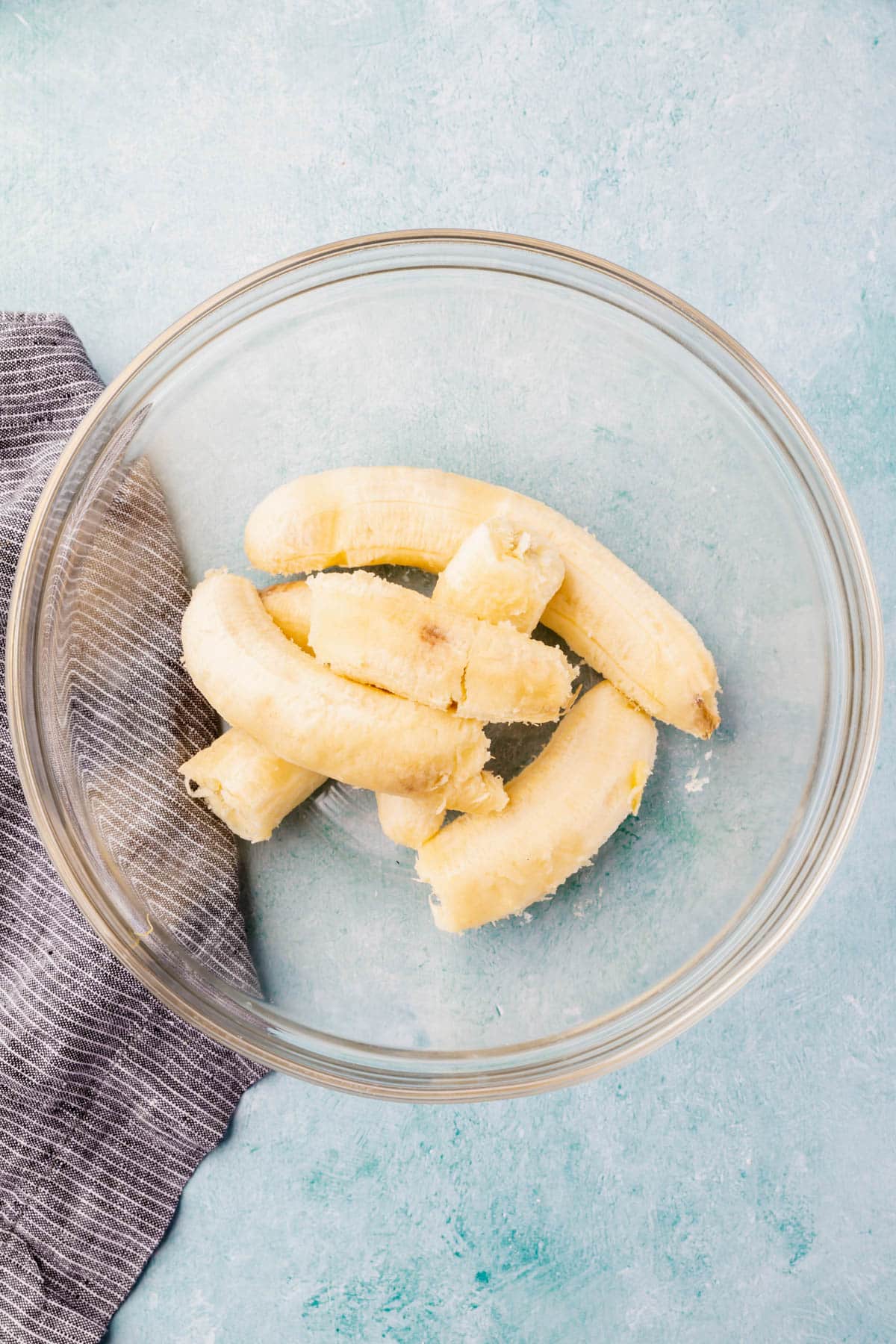 Ripe bananas in a large glass mixing bowl on a blue table.