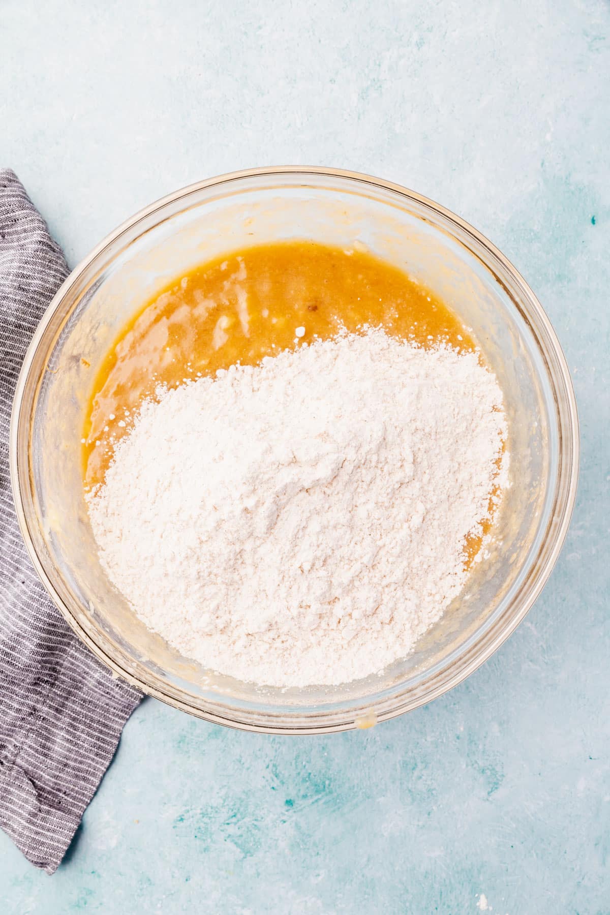 A glass mixing bowl with a wet banana mixture topped with gluten-free flour blend that has not yet been mixed in.