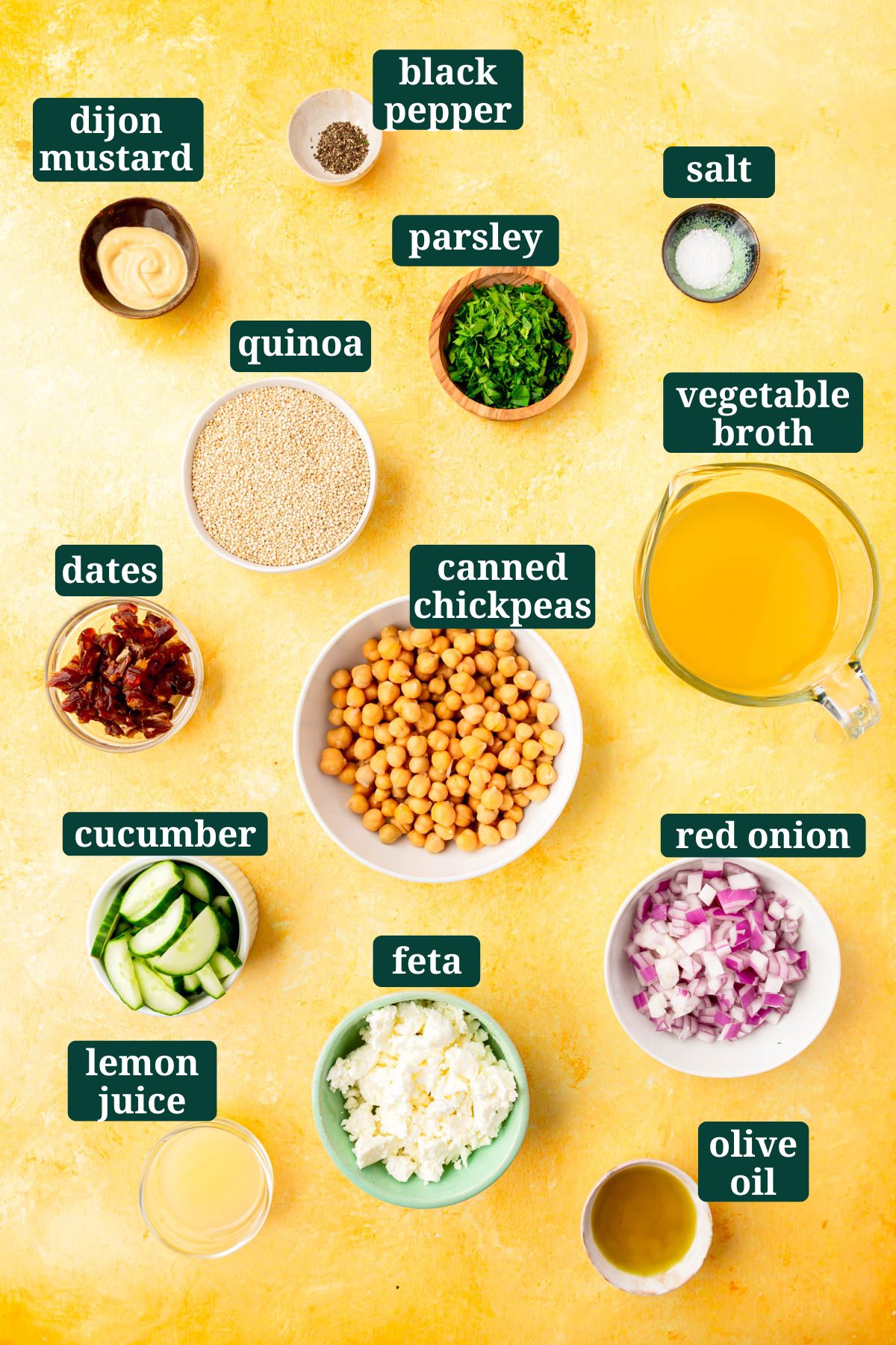 Ingredients in small bowls on a yellow table to make a cold quinoa chickpea salad, including fresh chopped parsley, dijon mustard, red onions, feta, cucumber, dates, salt, vegetable broth, and oil with text overlays over each ingredient.