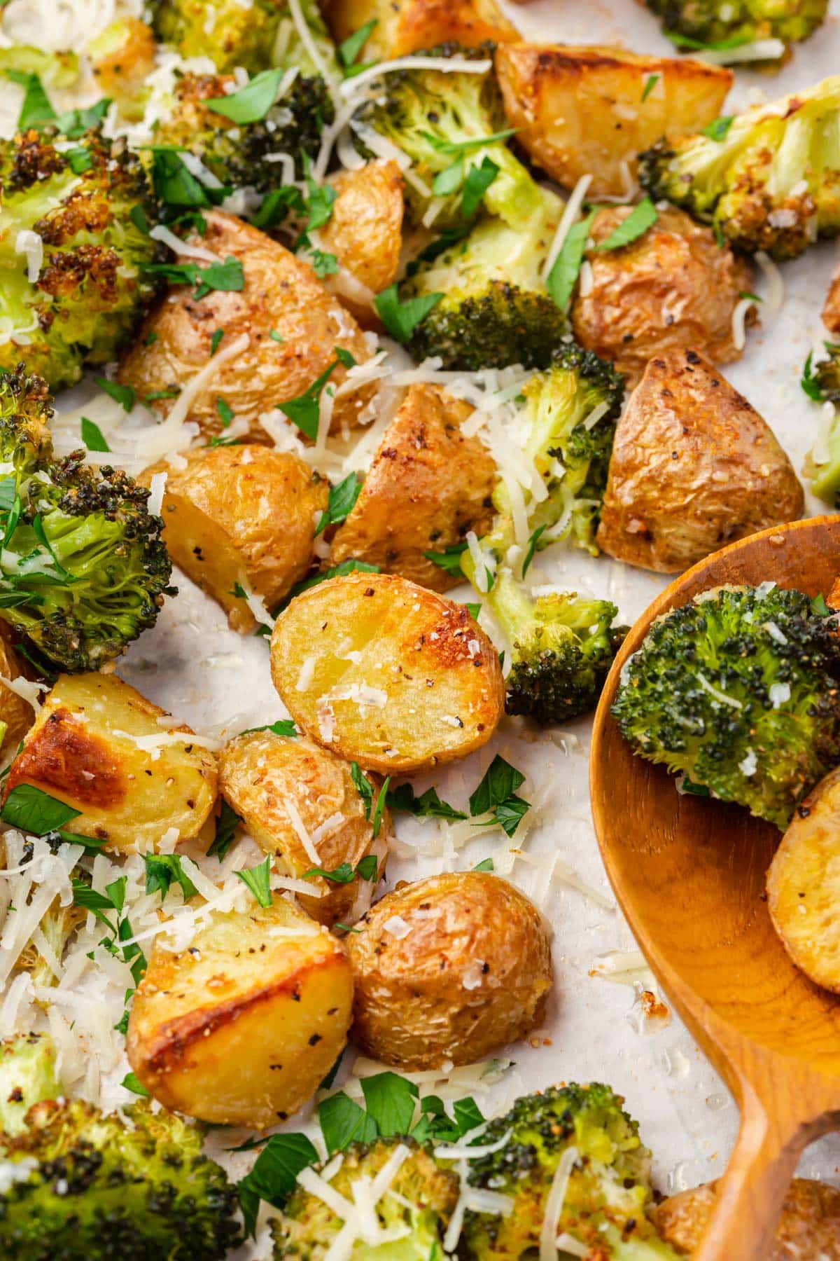 A closeup of roasted broccoli and potatoes topped with spices, shredded parmesan cheese, and parsley with a wooden serving spoon.