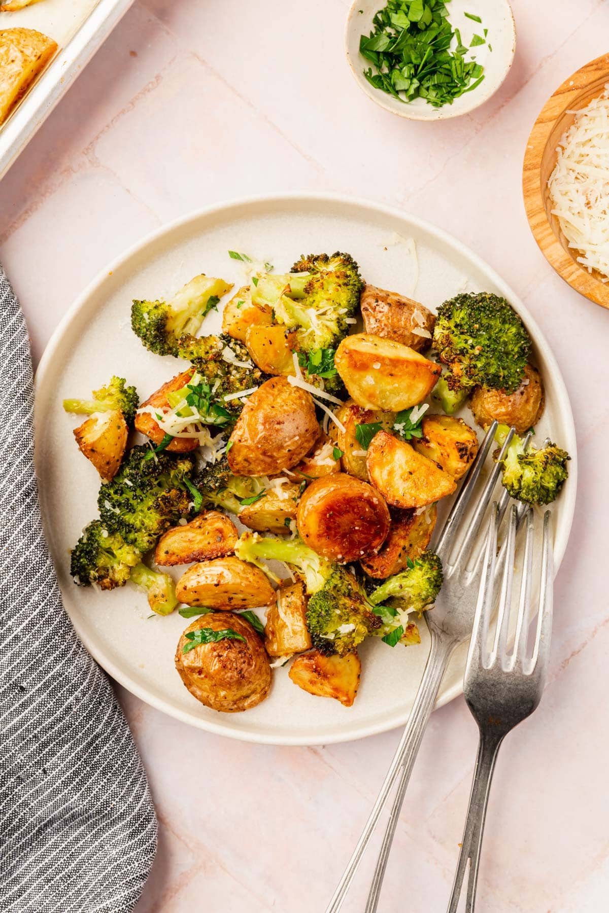 A plate filled with roasted baby potatoes and broccoli with two forks and a side of parmesan cheese and chopped parsley.