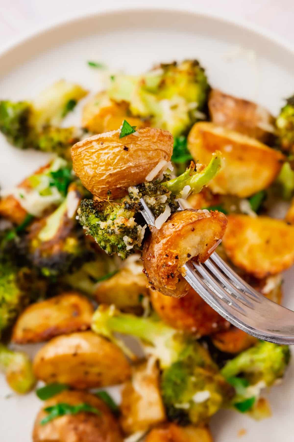 A fork holding a few pieces of roasted potatoes and broccoli topped with parmesan cheese over a bowl of roasted vegetables.