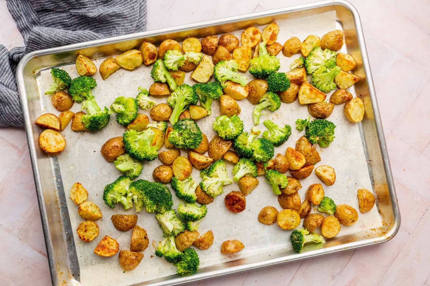 An overhead view of baby potatoes and broccoli on a baking sheet tossed together before baking.