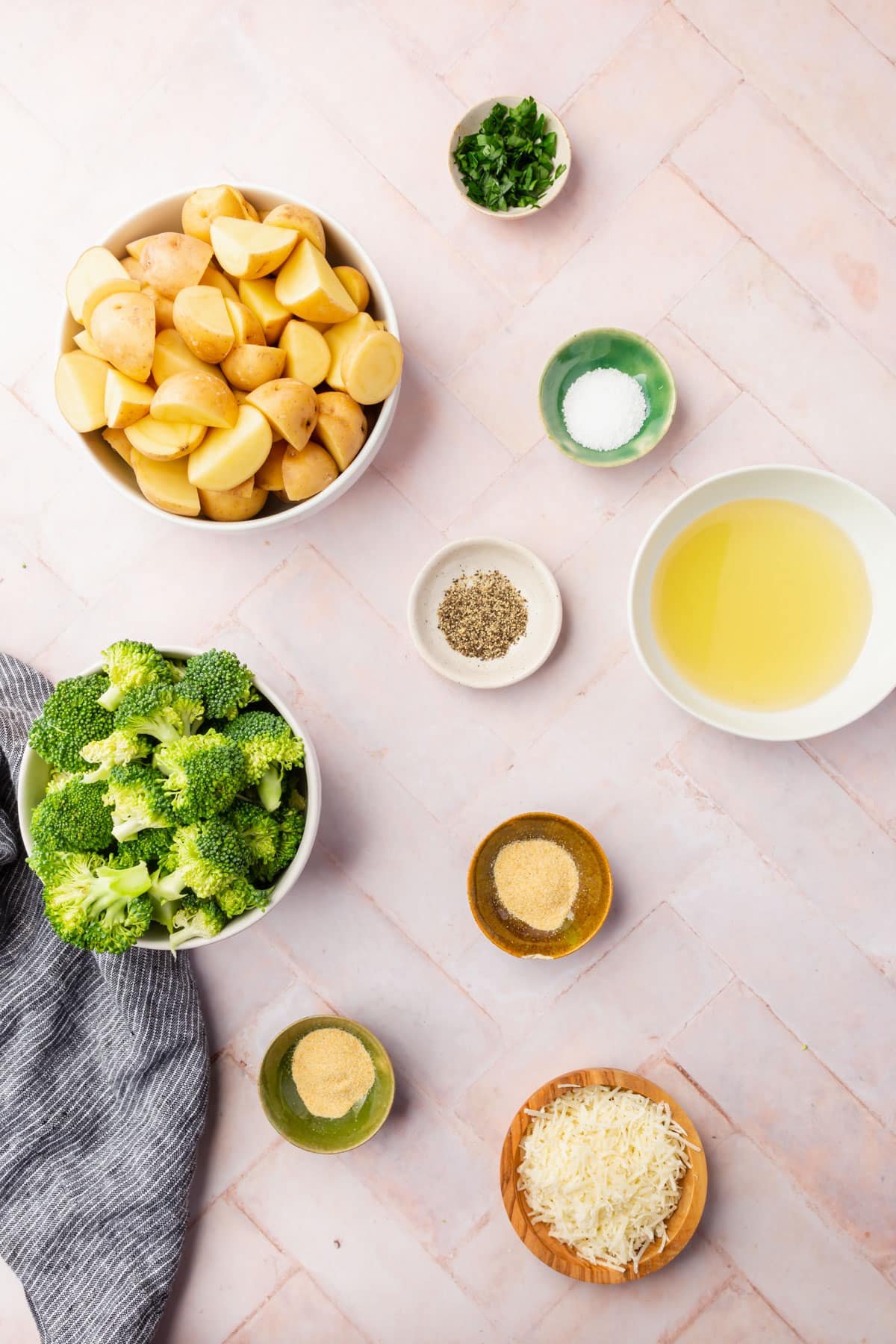 An overhead view of ingredients in bowls to make roasted broccoli ad potatoes, including baby potatoes, broccoli, parsley, pepper, salt, parmesan cheese, olive oil, garlic powder, and onion powder.