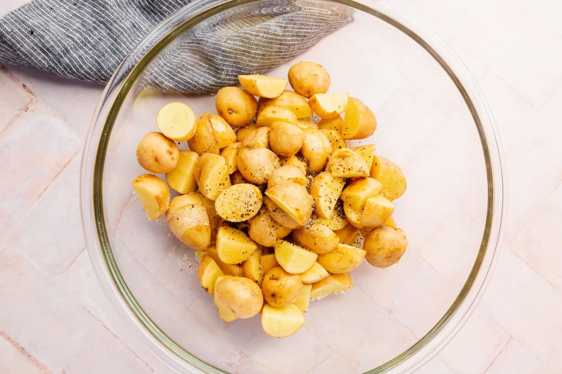 A glass bowl with sliced baby yukon gold potatoes topped with olive oil, salt, pepper, garlic powder and onion powder.