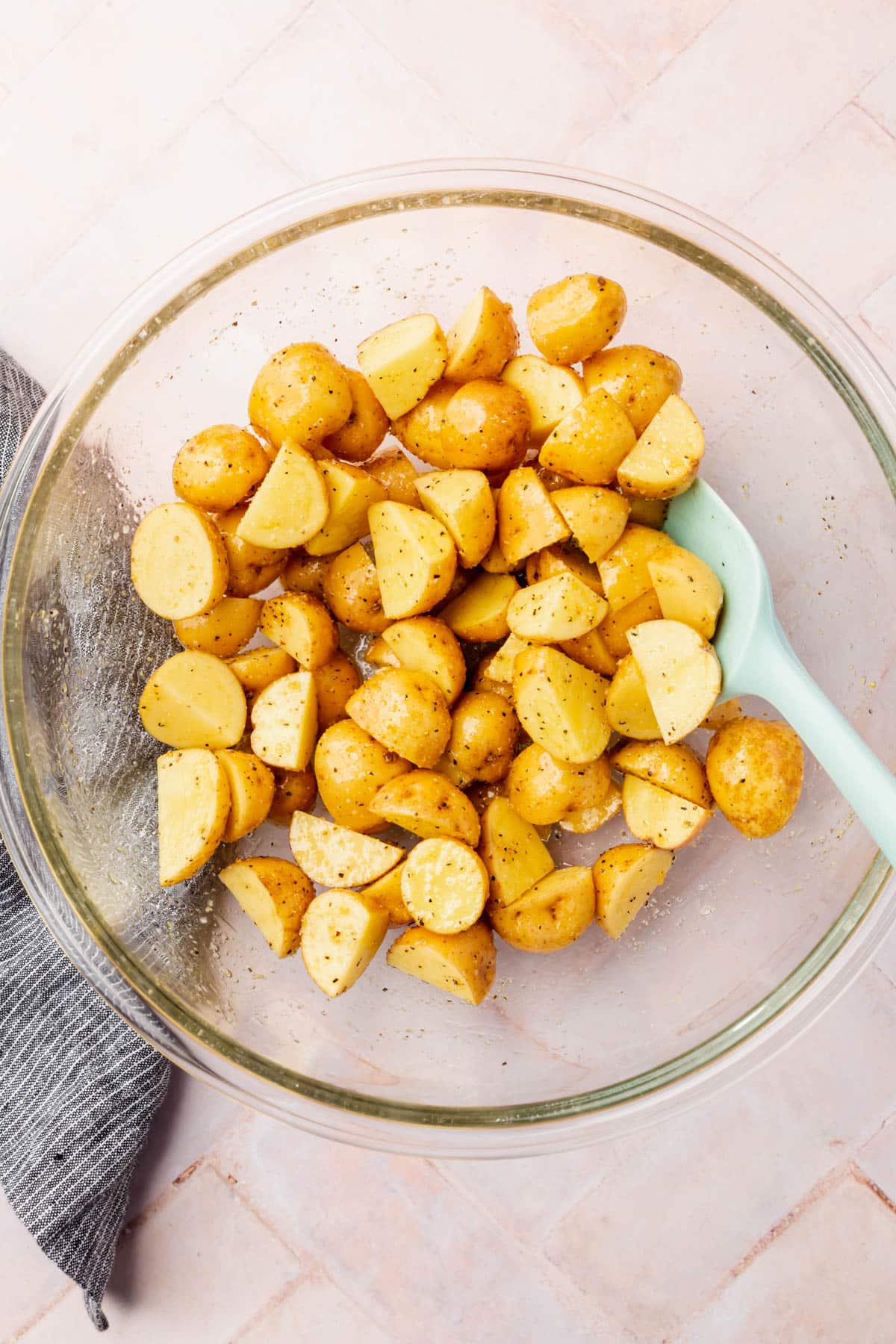 A glass mixing bowl of sliced baby yukon gold potatoes that have been mixed together with olive oil and spices with a blue spatula.