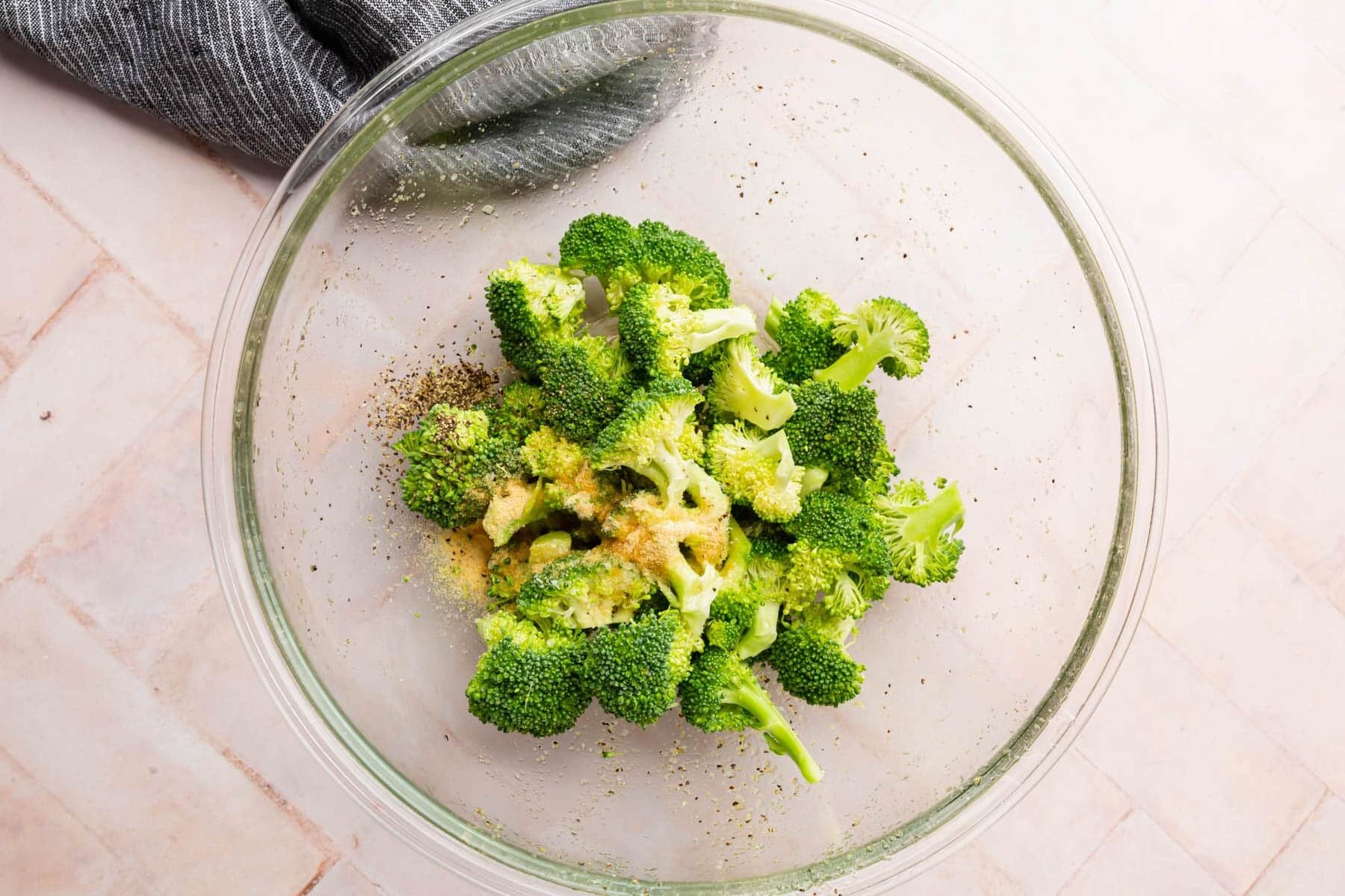 A glass mixing bowl with broccoli florets topped with olive oil, garlic powder, onion powder, salt, and pepper.