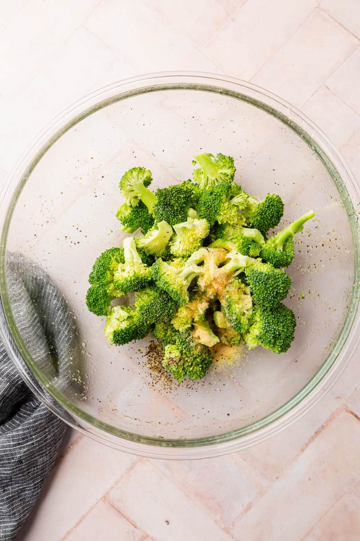 A glass mixing bowl with broccoli florets topped with olive oil, garlic powder, onion powder, salt, and pepper.