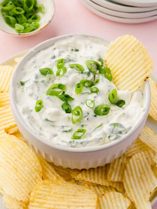 A bowl of green onion dip on a plate of Ruffle potato chips with a bowl of sliced green onions and a stack of plates in the background.