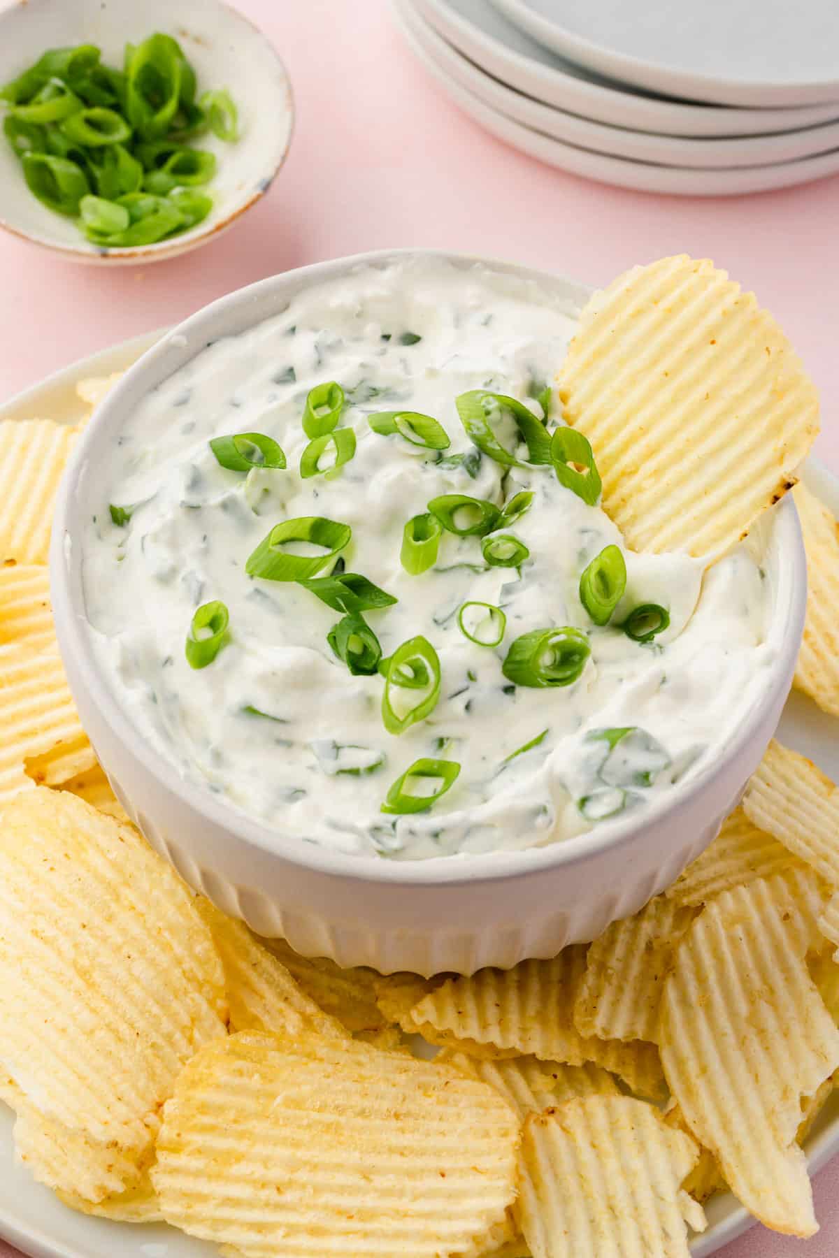 A bowl of green onion dip on a plate of Ruffle potato chips with a bowl of sliced green onions and a stack of plates in the background.