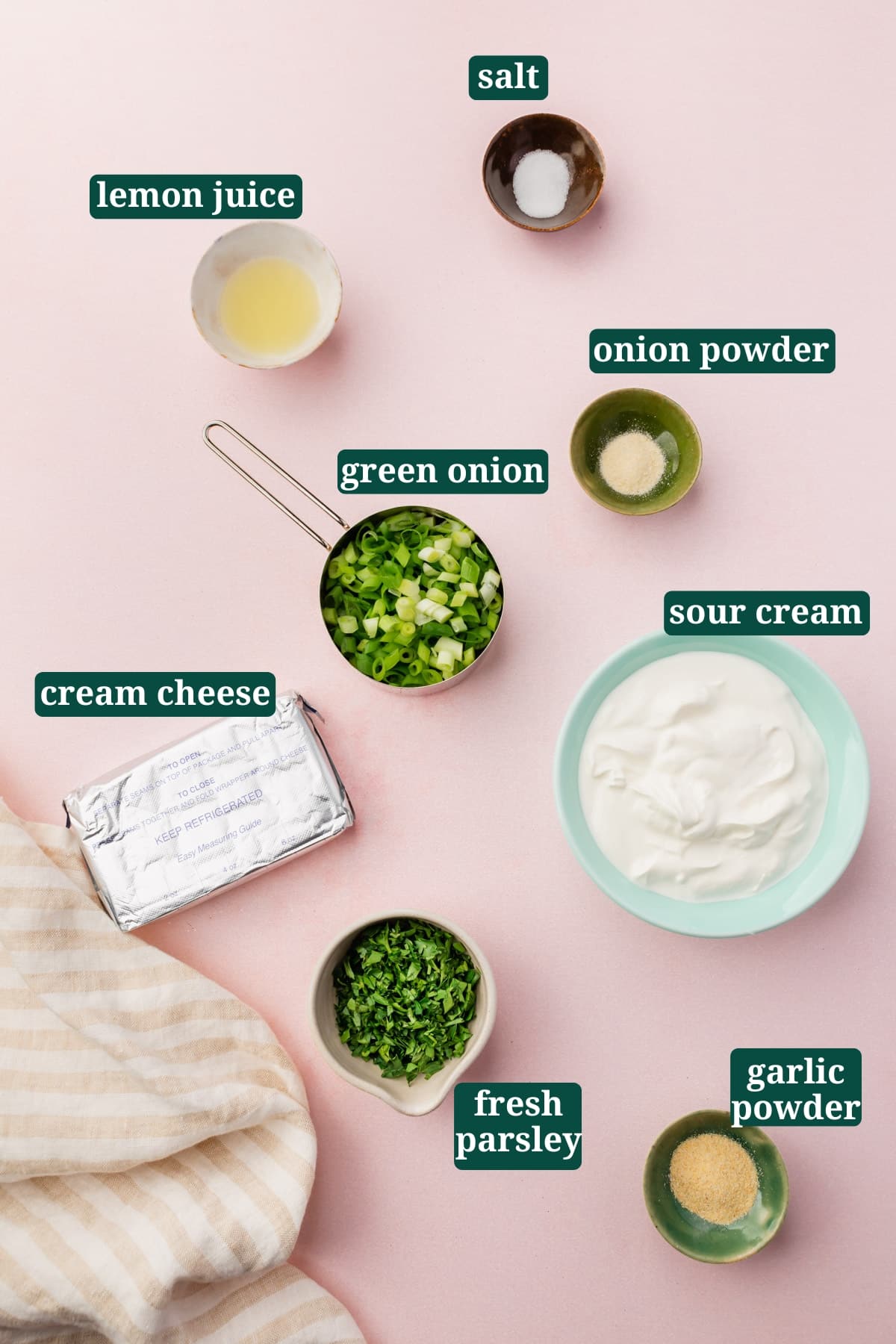 Small ingredients in bowls on a pink table for making green onion dip, including lemon juice, fresh parsley, green onions, cream cheese, sour cream, onion powder, garlic powder, and salt with a text overlay over each ingredient.