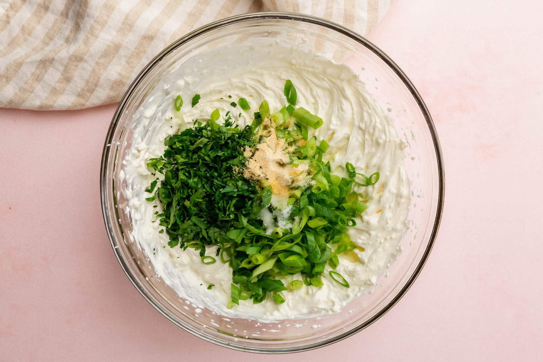 A glass mixing bowl with sour cream topped with sliced green onions, fresh parsley, garlic powder, onion powder, salt and lemon juice before mixing together.