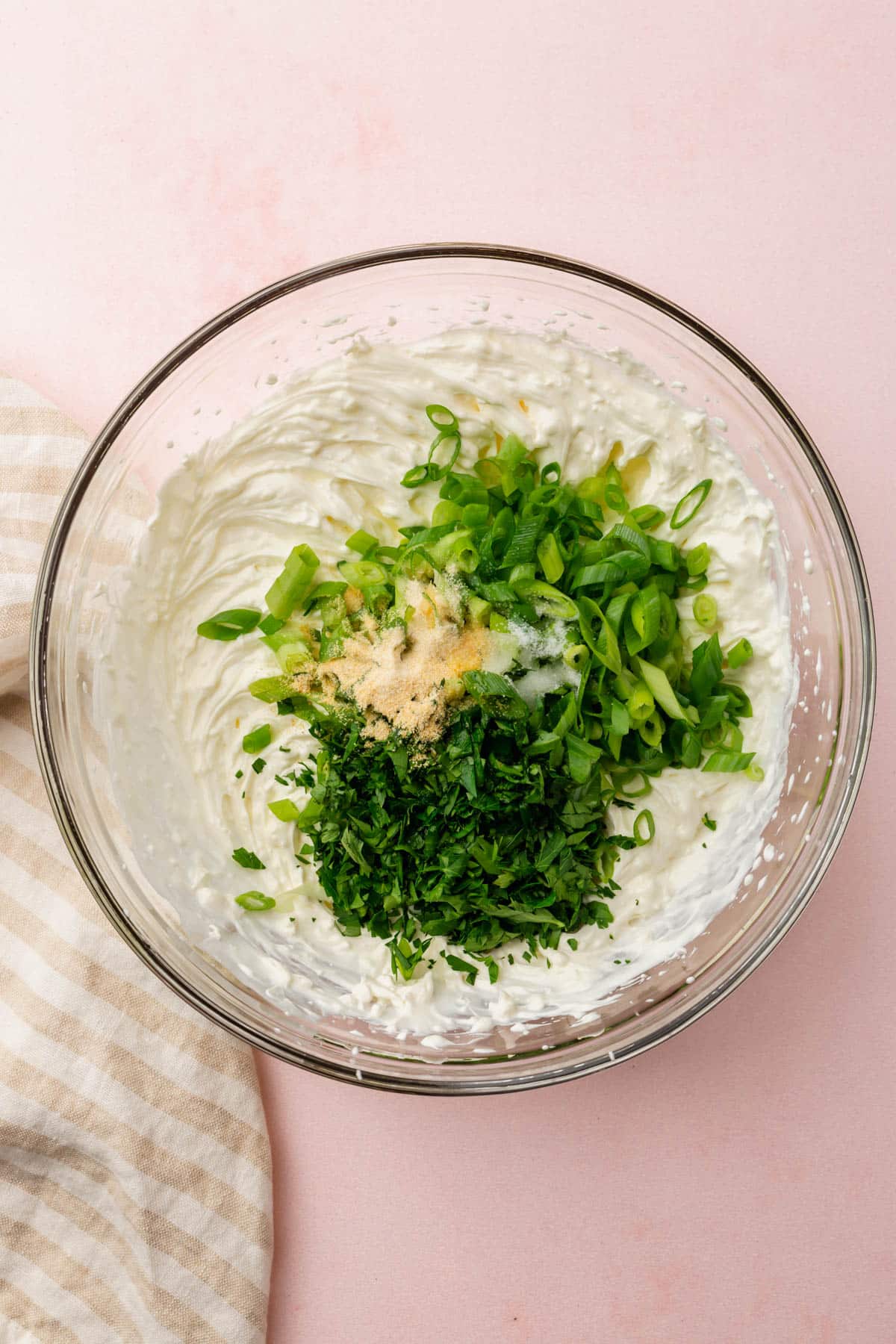 A glass mixing bowl with sour cream topped with sliced green onions, fresh parsley, garlic powder, onion powder, salt and lemon juice before mixing together.