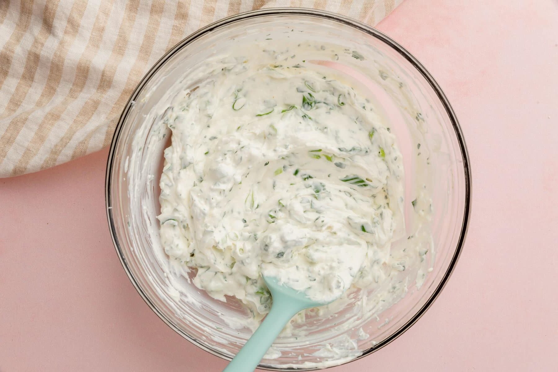 A glass mixing bowl of green onion dip with a blue spatula on a pink table.
