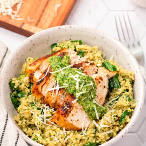 A bowl of spinach and quinoa topped with sliced chicken breast, parsley pesto and shredded parmesan cheese with a block of parmesan in the background that has been partially shredded.