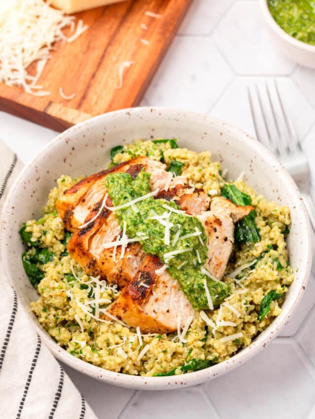 A bowl of spinach and quinoa topped with sliced chicken breast, parsley pesto and shredded parmesan cheese with a block of parmesan in the background that has been partially shredded.