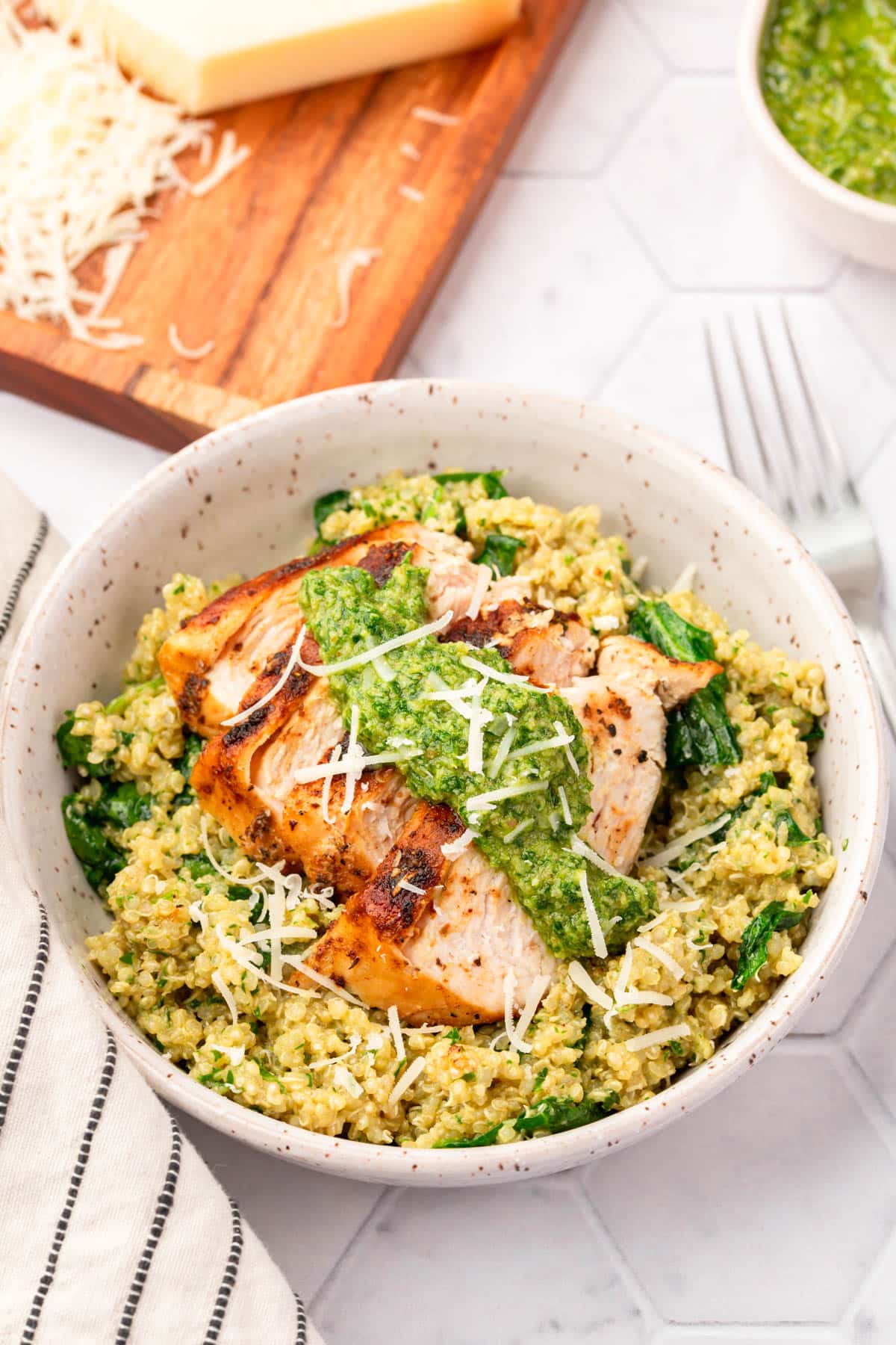 A bowl of spinach and quinoa topped with sliced chicken breast, parsley pesto and shredded parmesan cheese with a block of parmesan in the background that has been partially shredded.