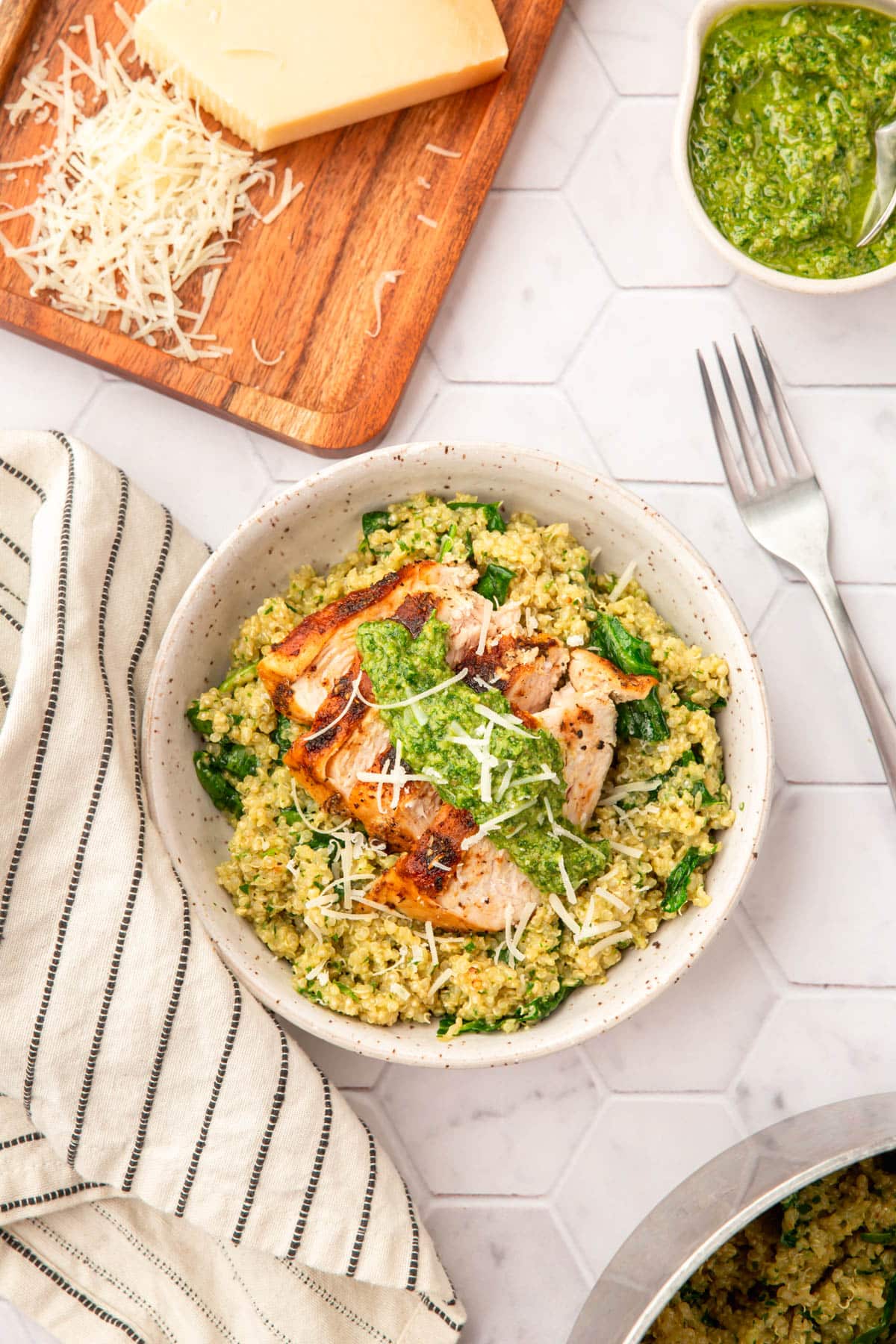 A bowl of pesto chicken on top of a bed of spinach quinoa with a fork, a bowl of parsley pesto, a block of parmesan cheese that has been partially shredded.