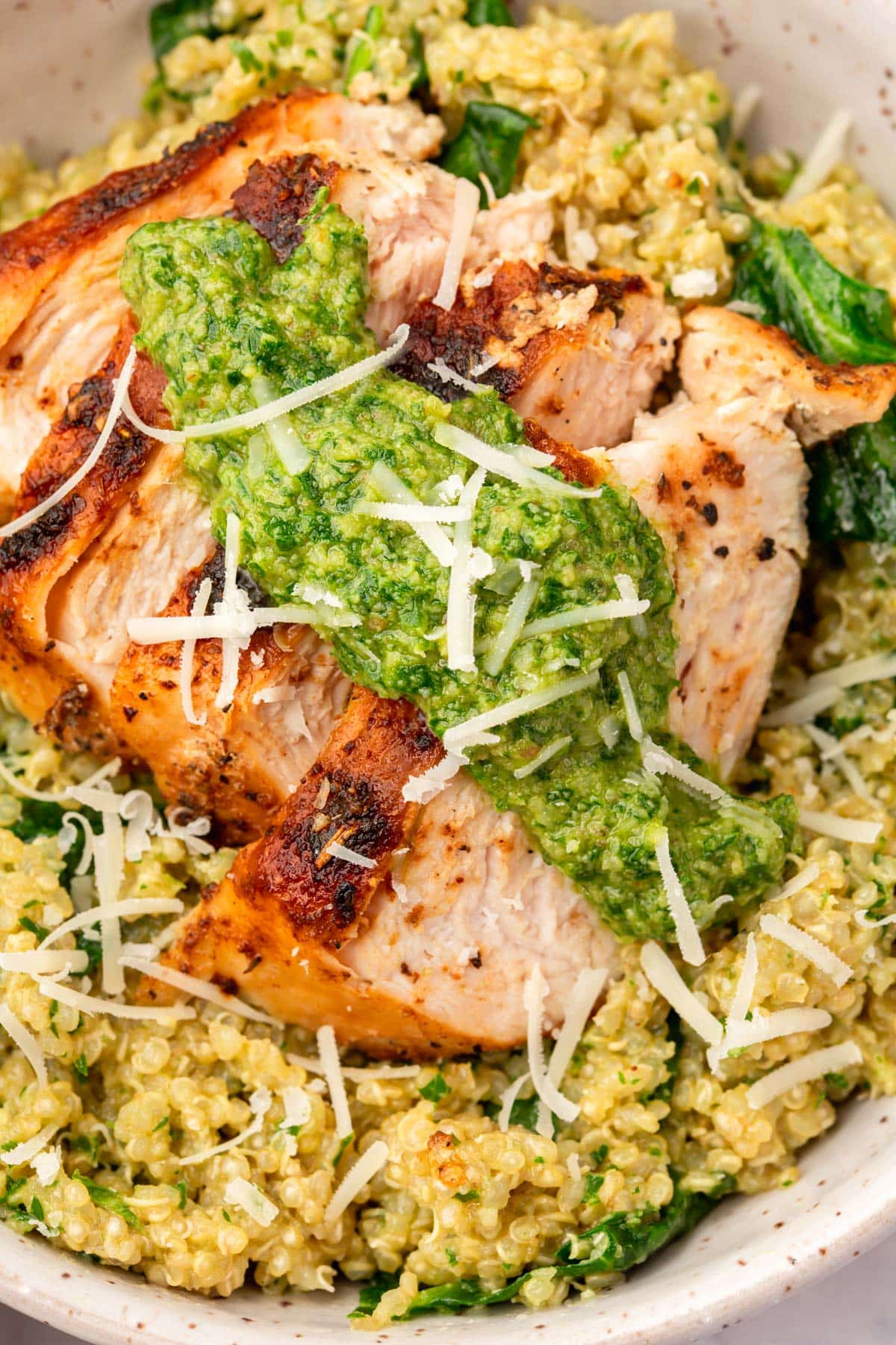 A close up of a bowl of quinoa mixed with spinach and topped with sliced chicken breast, parsley pesto and shredded parmesan cheese.