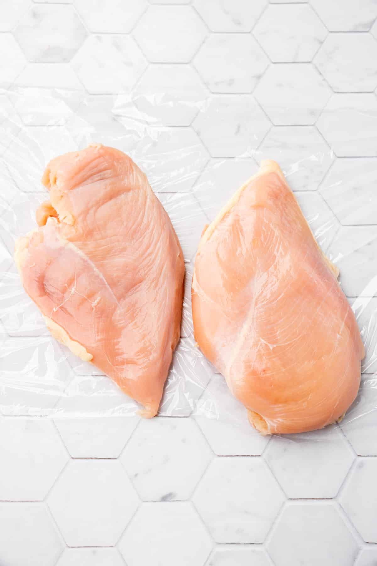 Two raw chicken breasts on a piece of plastic wrap.