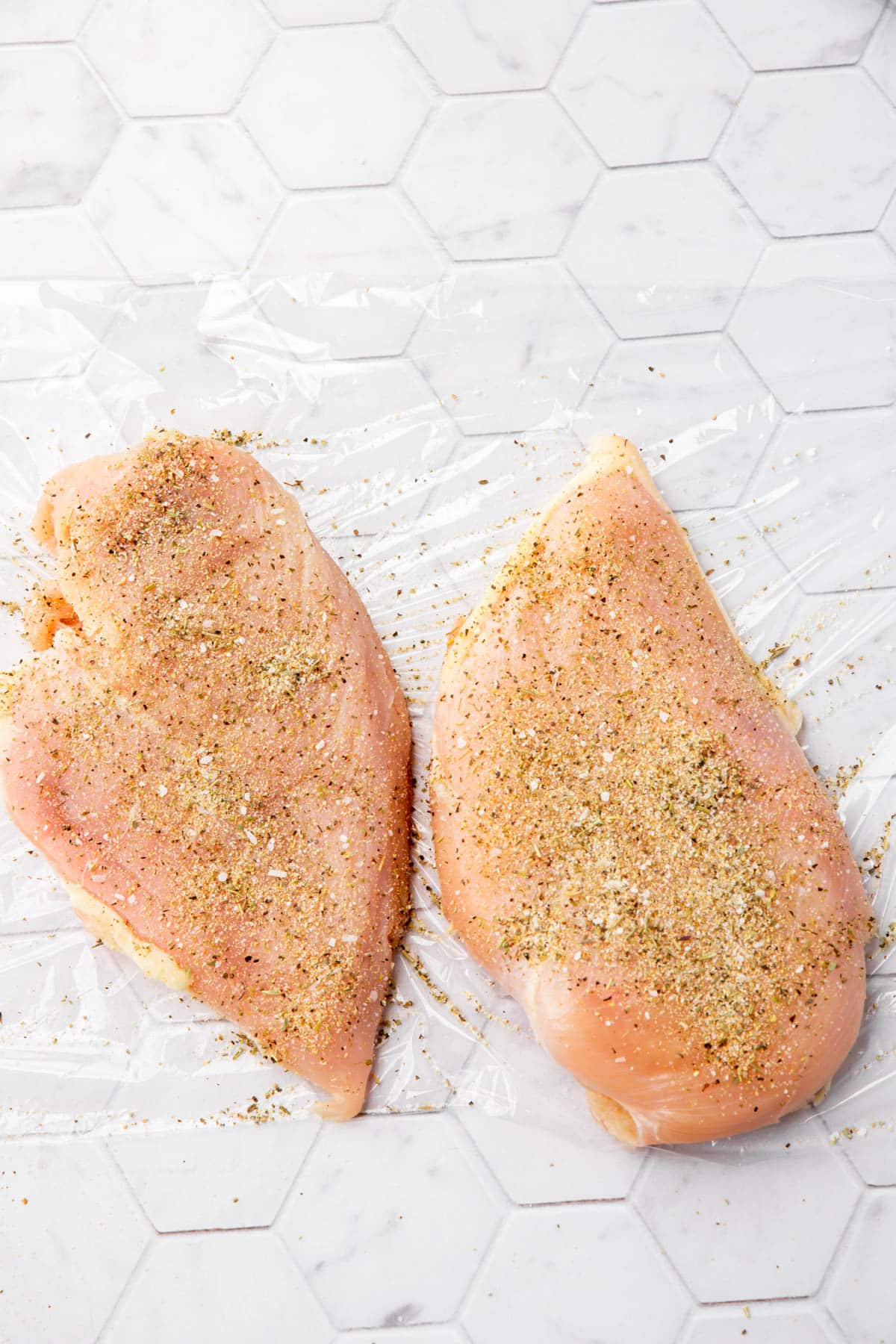 Two raw chicken breasts on a piece of plastic wrap that have been topped with spices.