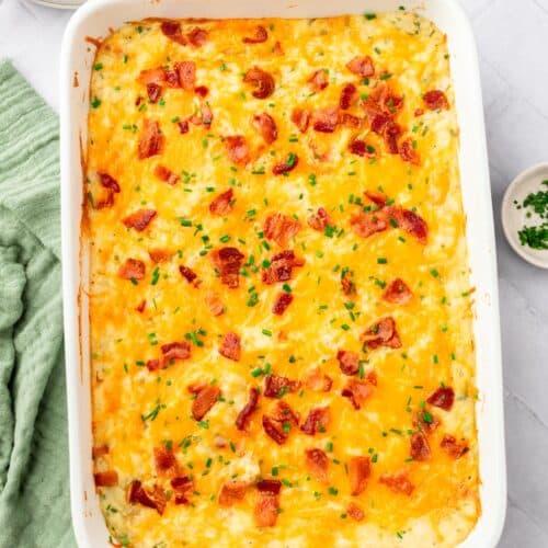 A casserole dish of twice baked mashed potatoes topped with bacon bits, chopped chives and melted cheddar cheese.