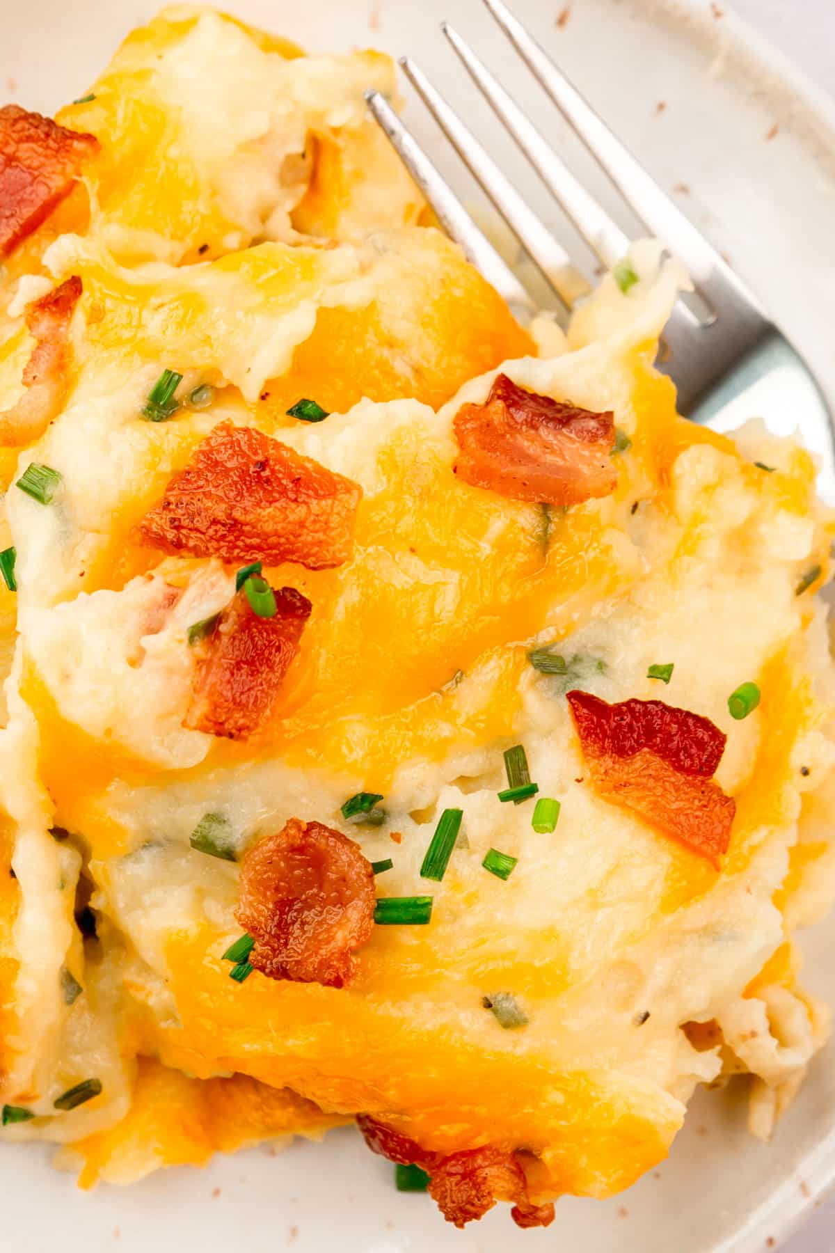 A closeup of a plate of twice baked mashed potatoes topped with bacon bits, chopped chives and melted cheddar cheese with a fork next to it.