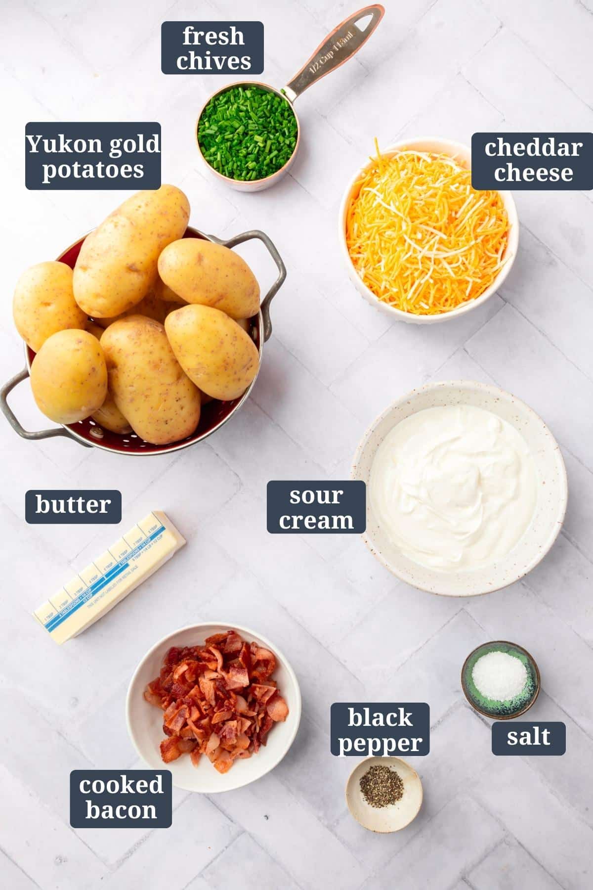 Ingredients in bowls on a gray chevron table to make twice baked mashed potatoes, including yukon gold potatoes, chives, cheddar cheese, butter, sour cream, cooked bacon bites, black pepper and salt with text overlays over each ingredient.