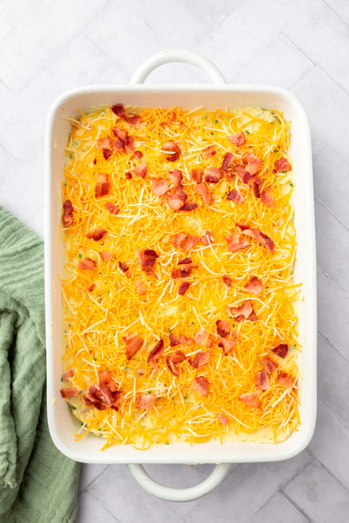 A casserole dish filled with mashed potatoes topped with chopped bacon and shredded cheddar cheese before baking in the oven.