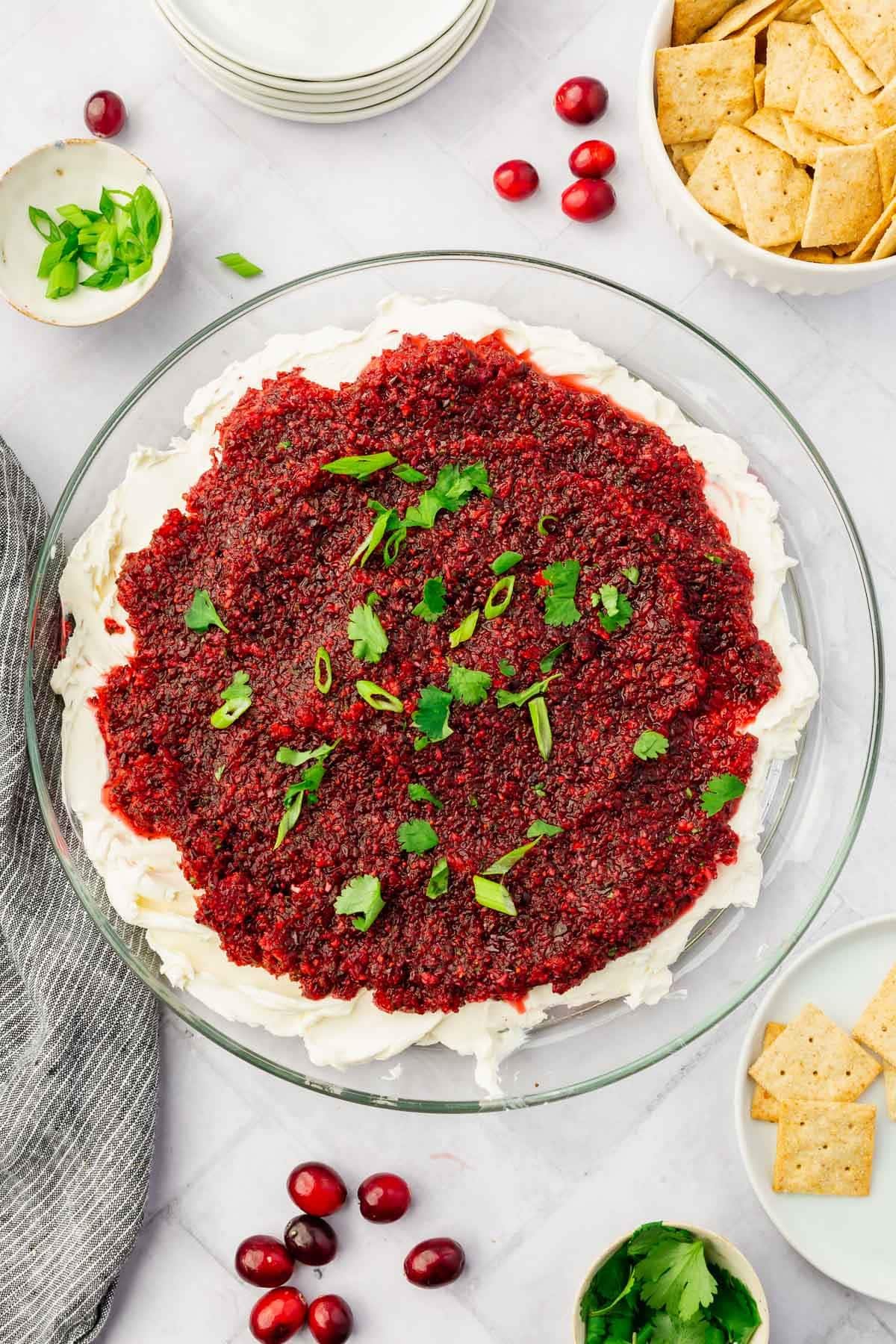 A platter of cream cheese topped with cranberry salsa, green onion and cilantro with gluten-free crackers on the side.