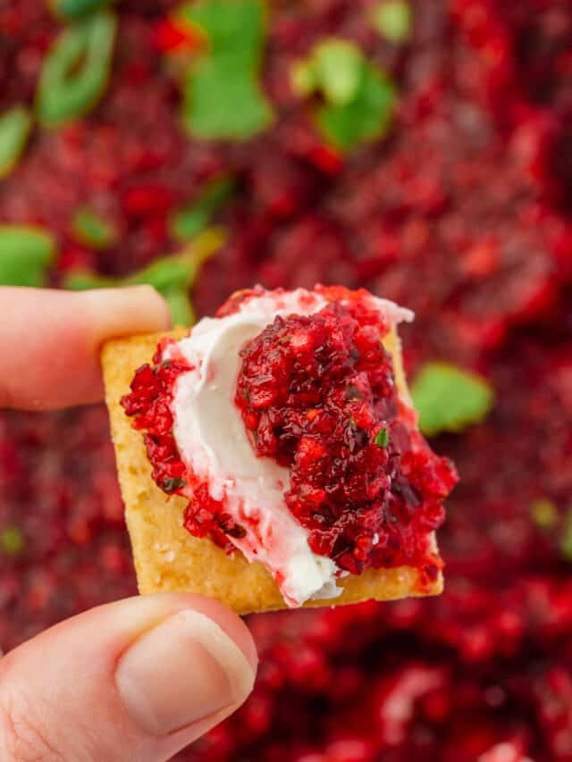 A hand holding a gluten-free cracker topped with cream cheese and cranberry jalapeño salsa.