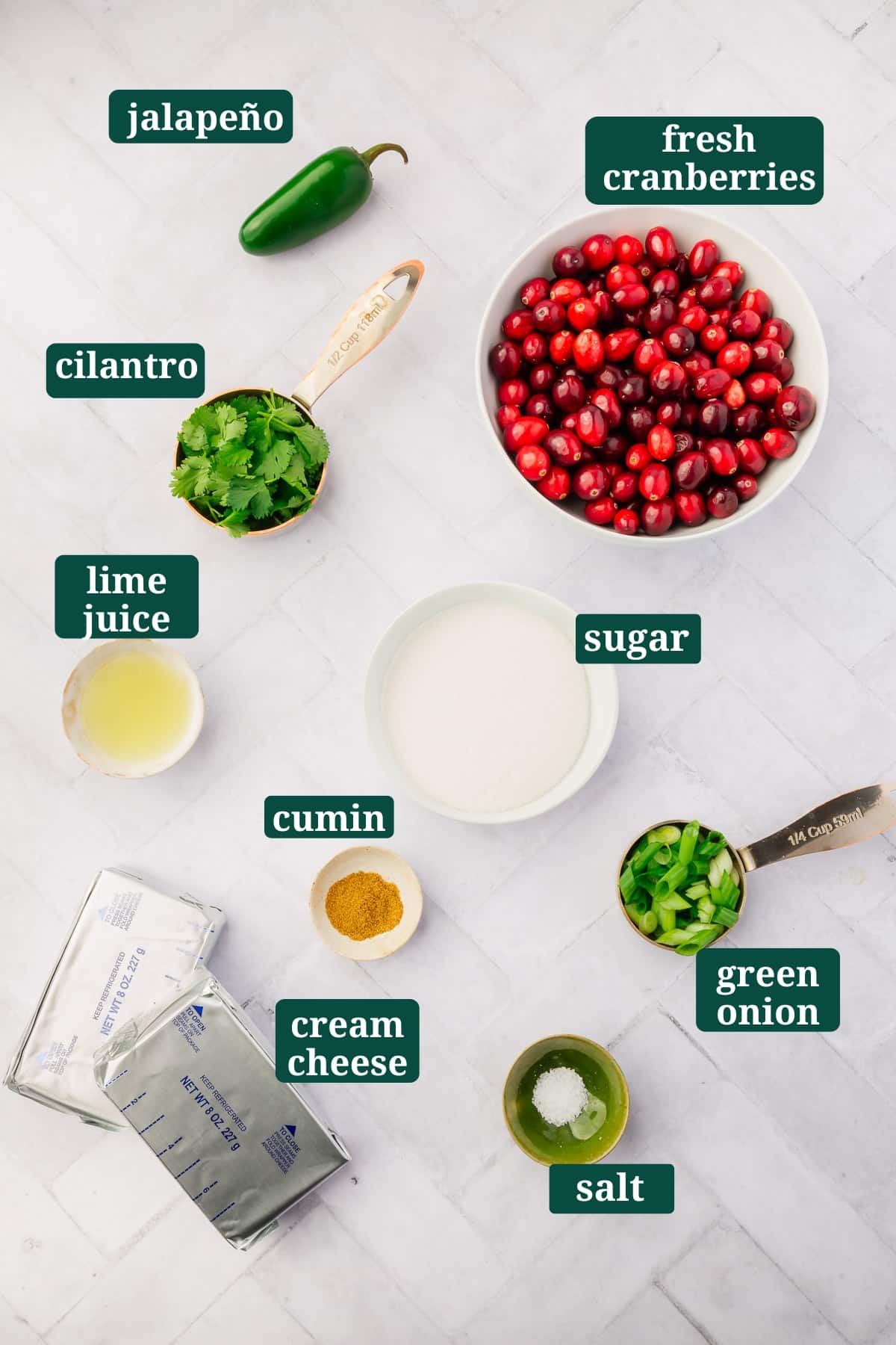Ingredients in small bowls to make cranberry cream cheese dip, including jalapeño, cranberries, green onions, sugar, lime juice, salt, and cilantro with text overlays over each ingredient.
