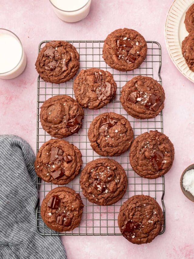 A cooling rack on a pink table filled with gluten-free chocolate chunk cookies topped with flaky sea salt with glasses of milk on the side.